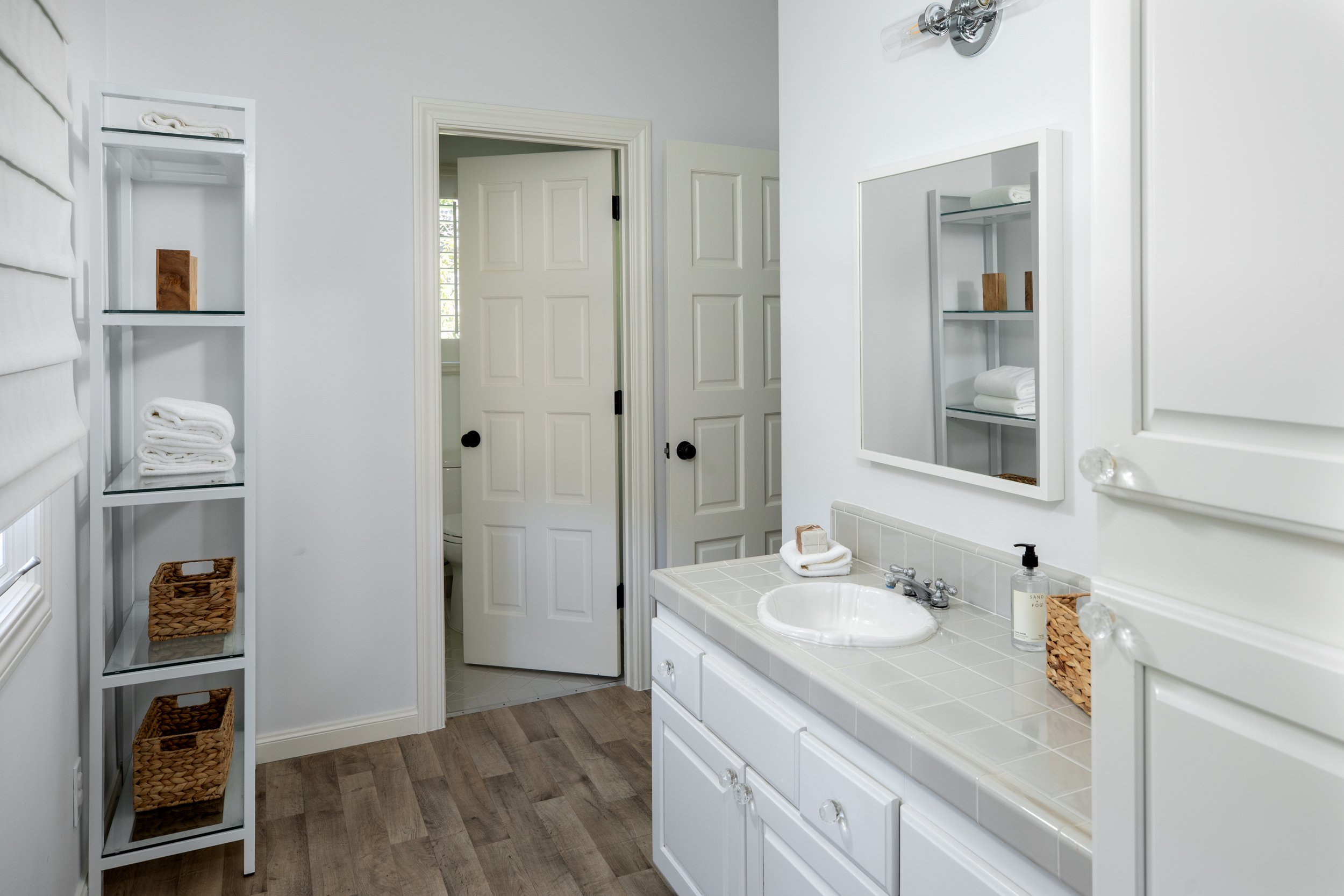 A white bathroom with a vanity, mirror, and shelves holding towels and baskets. Open door to a neighboring room with a window.