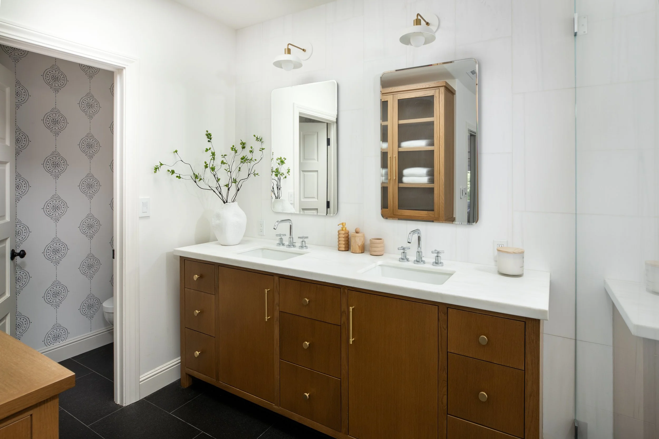 Modern bathroom with a white vanity, wooden cabinet, large mirror, and two wall-mounted lights. Decor includes a white vase with green branches and neutral-colored containers. The space features white walls, black floor tiles, and wallpaper with blac