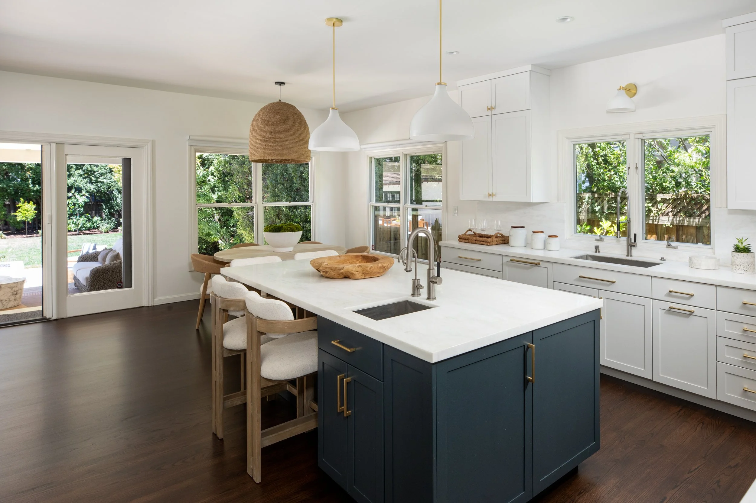 Modern kitchen with white cabinets, a dark blue island, pendant lights, windows showing a green outdoor yard, and a dining area with chairs and a view of the yard.
