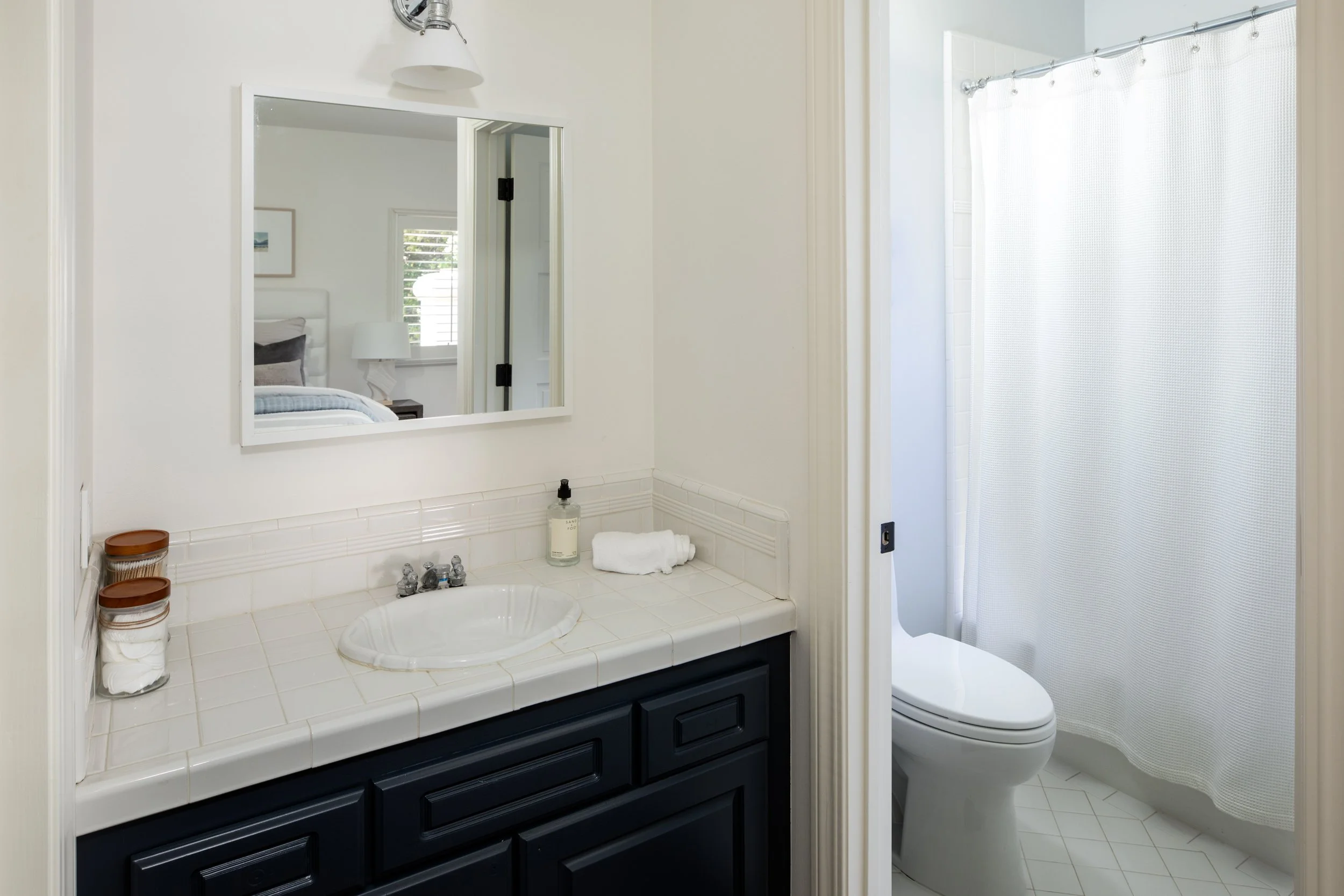 A bathroom with a white tiled vanity, black cabinet, and a mirror showing part of a bed and window. A bottle of hand soap, rolled towels, and small containers are on the vanity. To the right is a toilet and a shower with a white curtain.