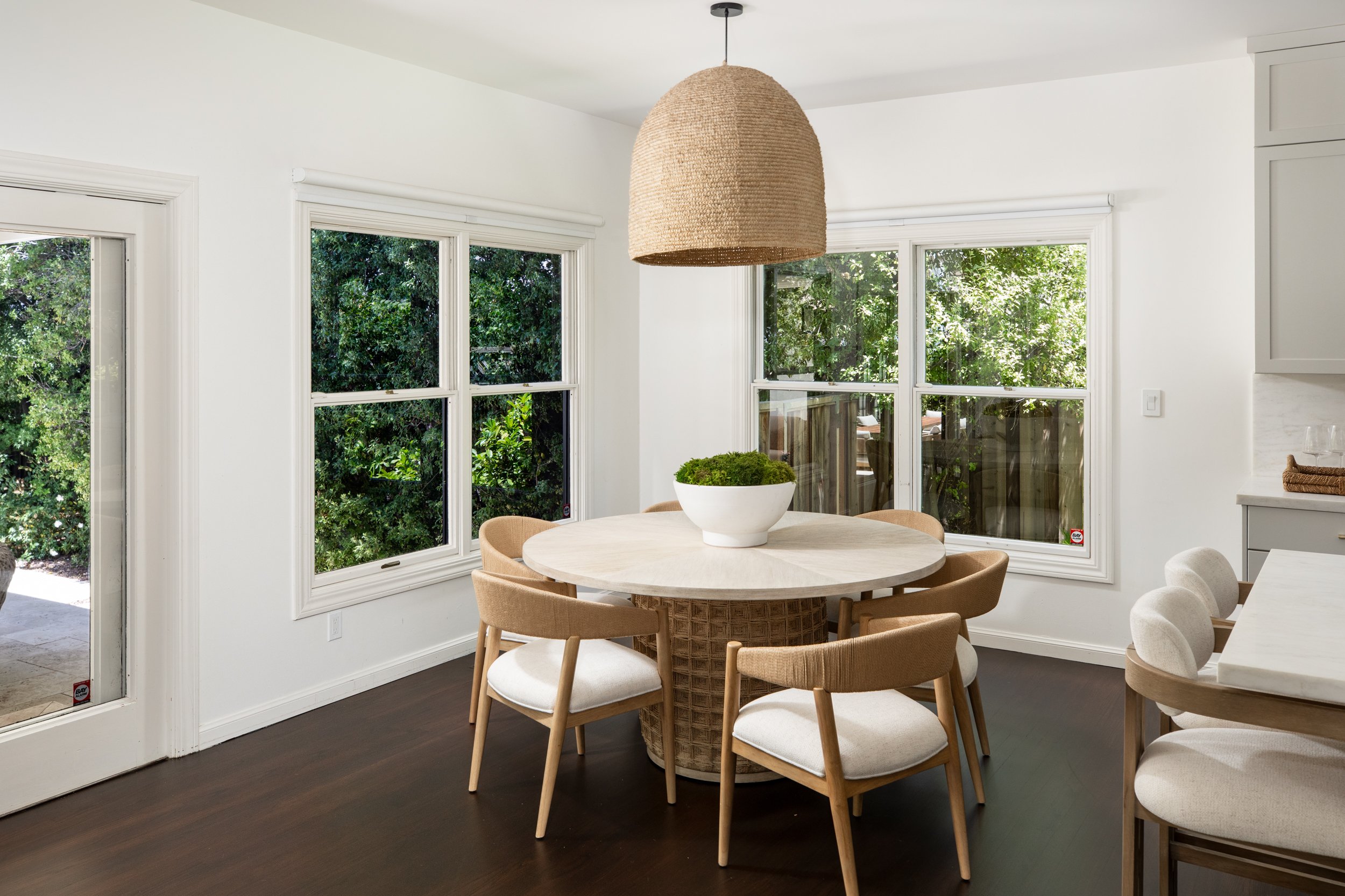 Bright dining area with white walls, dark hardwood floor, round marble-top table, and beige upholstered chairs. Large windows with white trim reveal lush green foliage outside. A woven pendant light hangs above the table, and a white bowl with greene