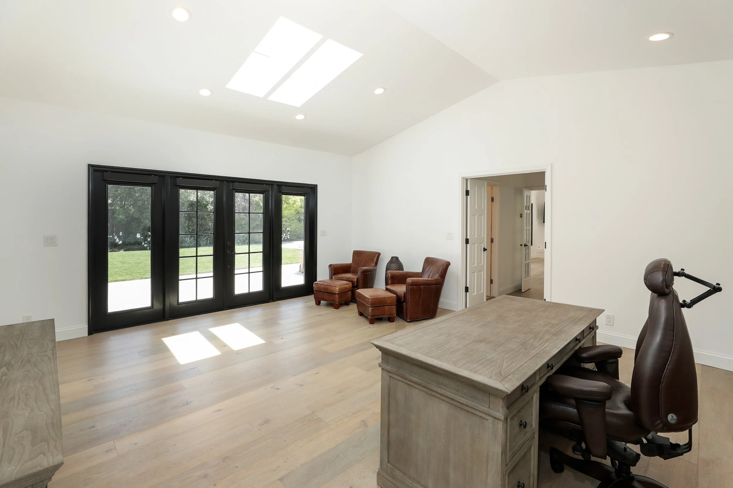 Living room with large black-framed glass double doors, white walls, wooden flooring, and a ceiling skylight. There are two brown armchairs with ottomans, a desk, and a rolling office chair.