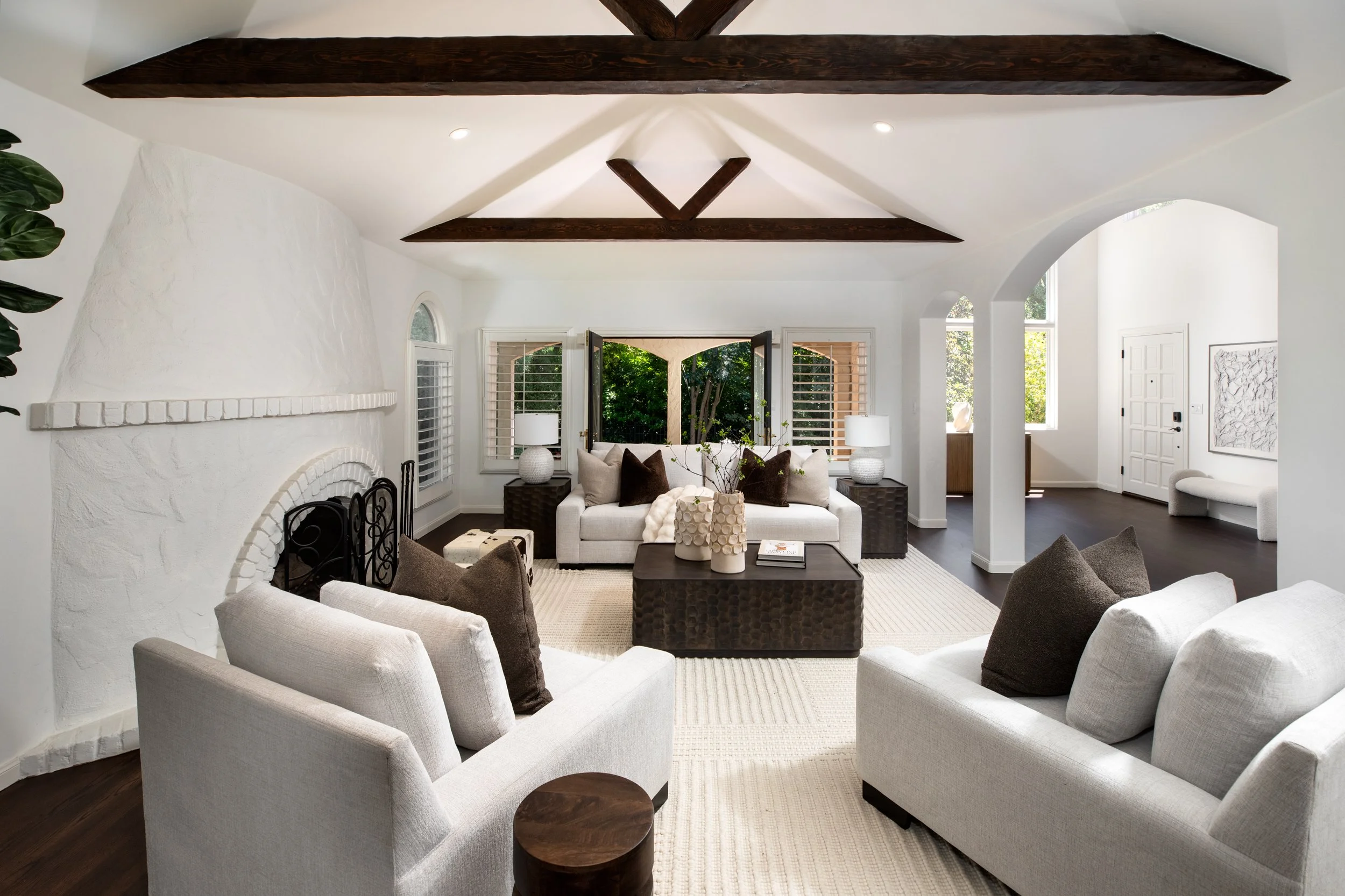 Living room with white sofas, dark pillows, a central dark wooden coffee table, and a white brick fireplace. Large windows with white plantation shutters and a view of green trees outside. Dark wooden beams on the ceiling.