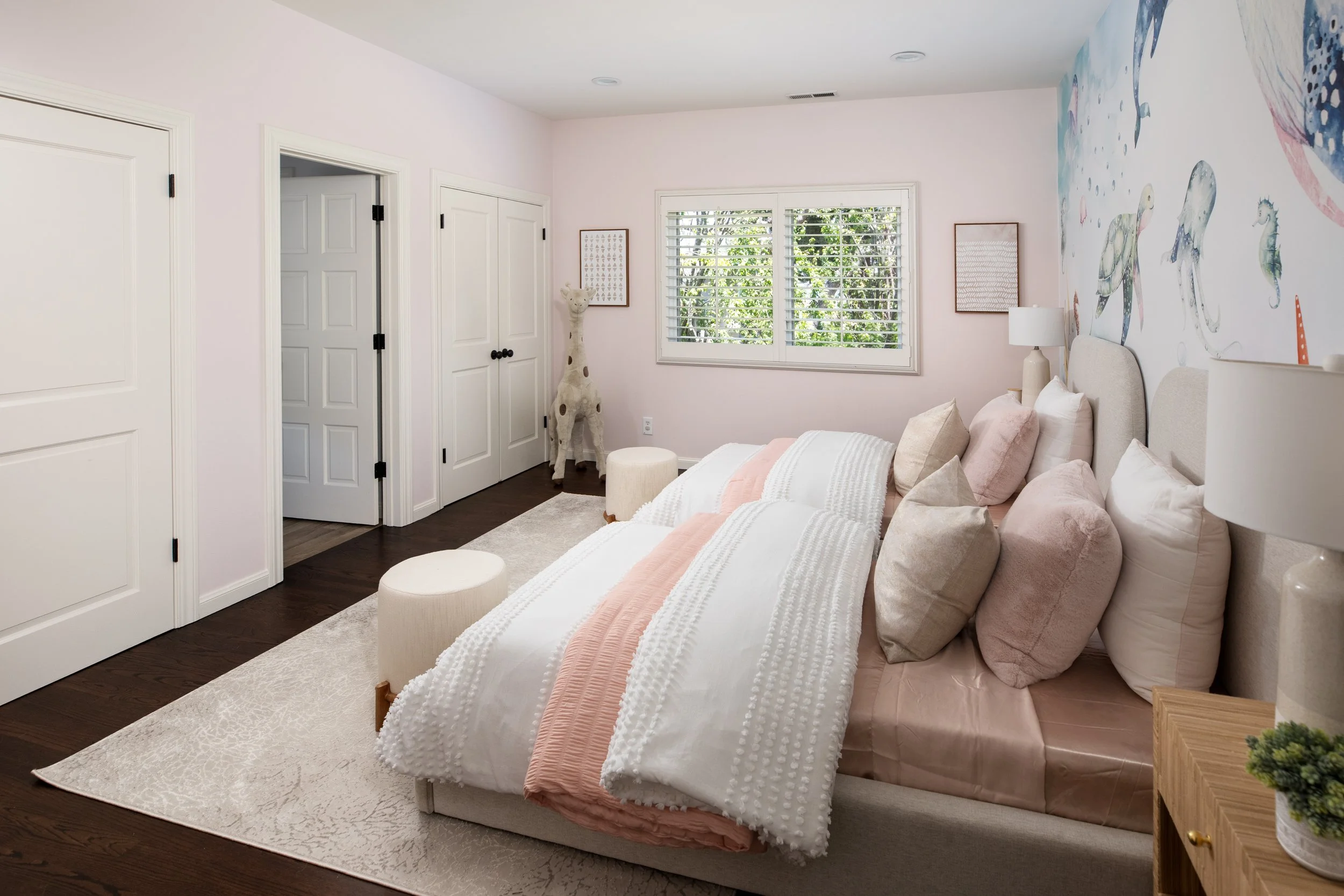 A cozy bedroom with three beds, each decorated with white and pink bedding and multiple pillows, a nightstand with a lamp, a large window with blinds, and a giraffe sculpture in the corner.