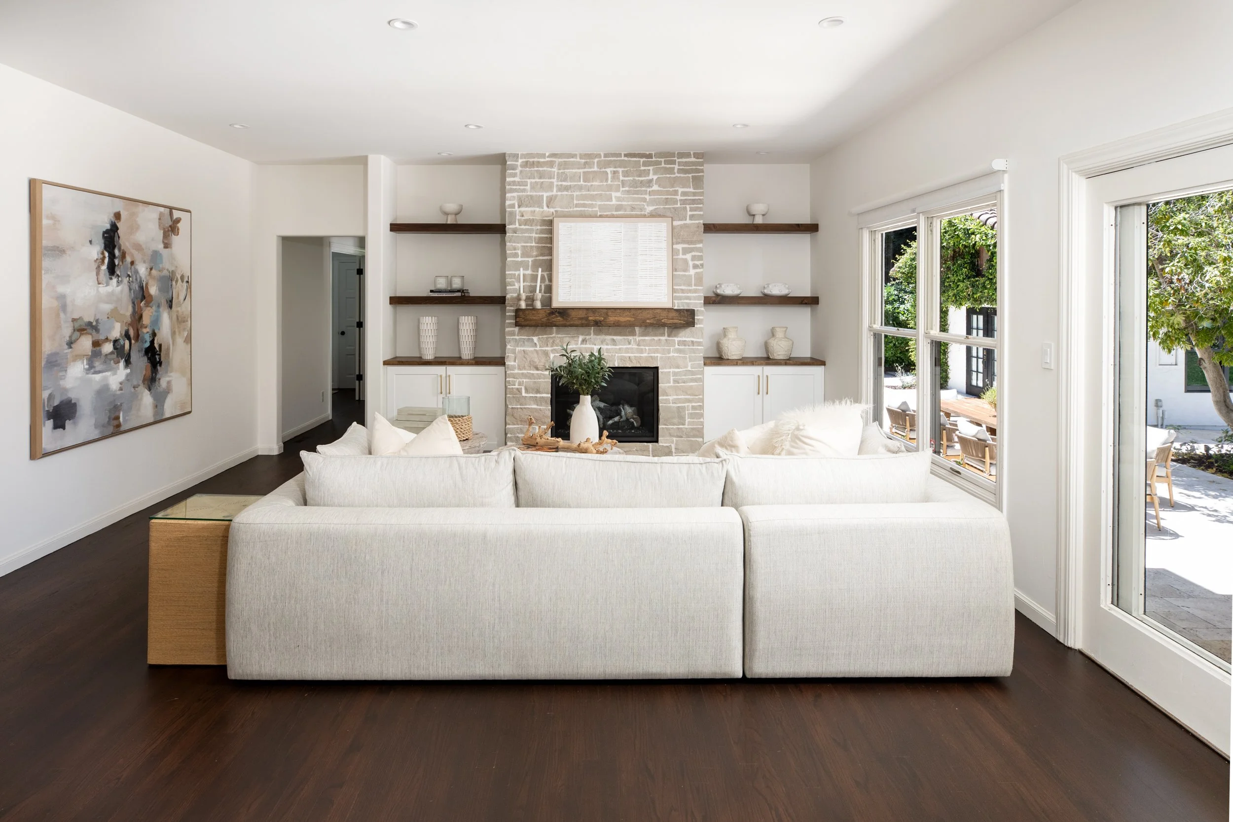 Living room with white sofa, dark wood floor, brick fireplace,floating shelves with decor, large window with outdoor seating.