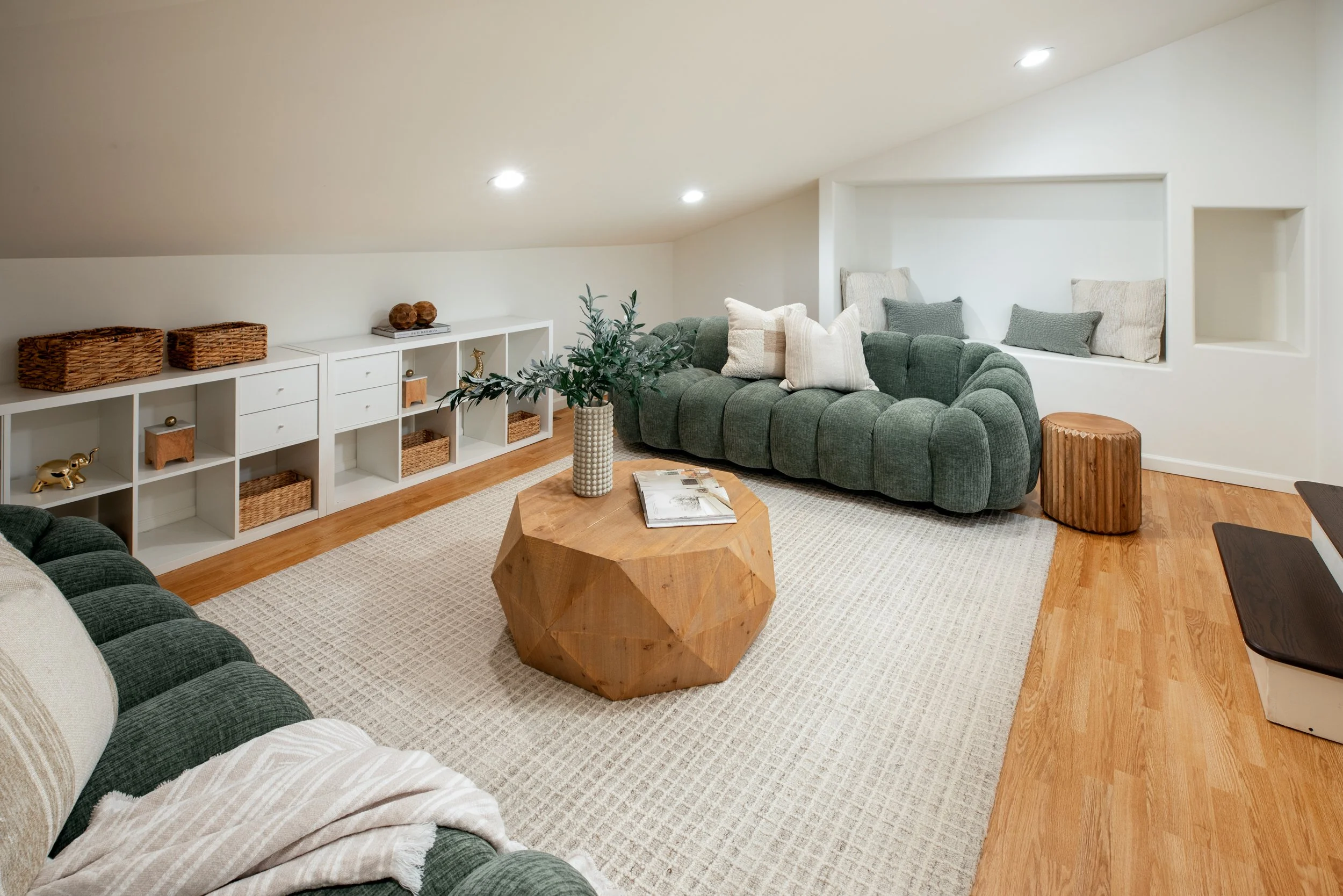 A cozy living room with a white ceiling, recessed lighting, a green tufted sectional sofa with pillows, a wooden coffee table with a plant and magazines, a white bookshelf with baskets and decor, a wicker stool, and a built-in window seat with cushio