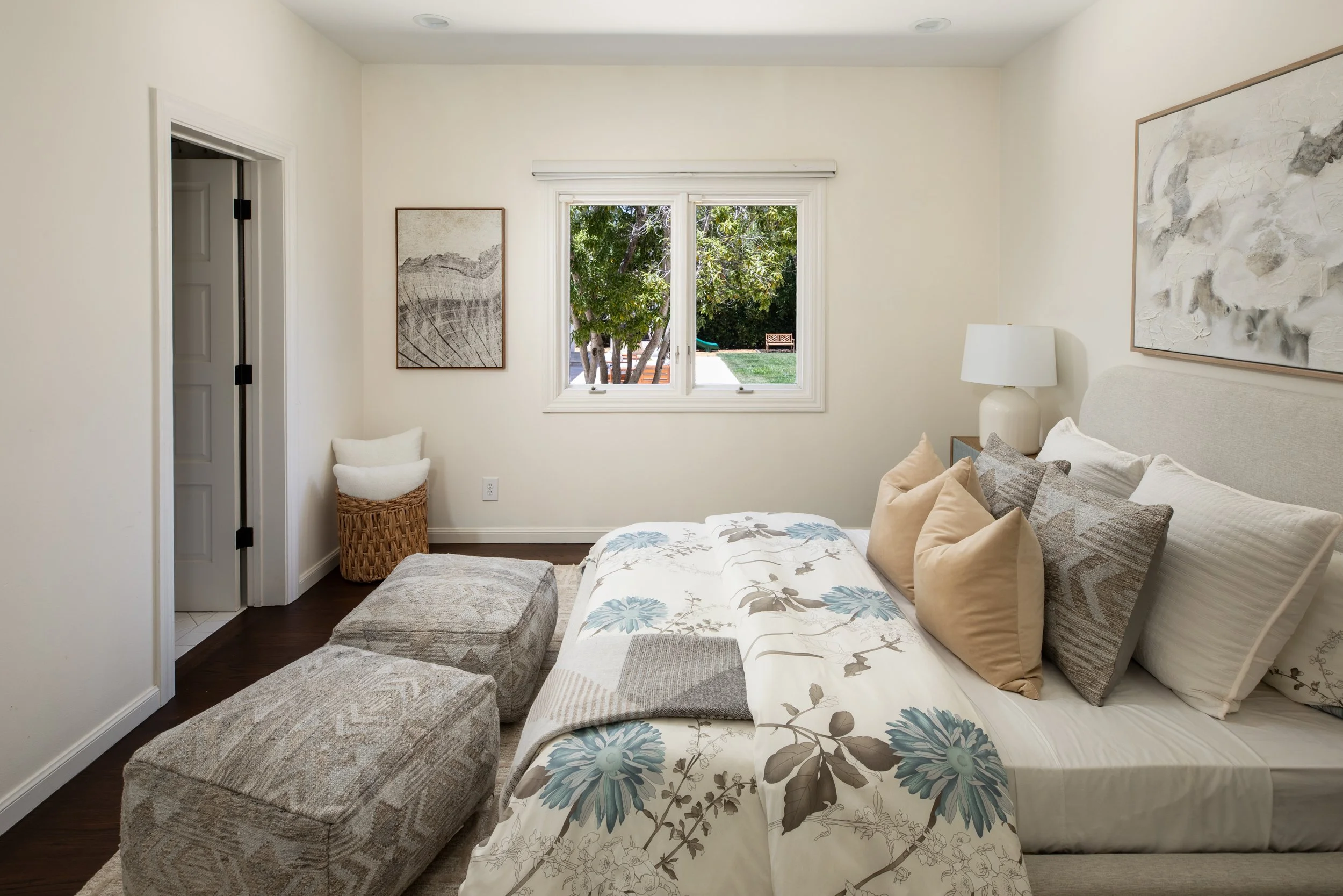 A bedroom with a bed, colorful pillows, painting on the wall, window overlooking trees and yard, nightstand with lamp, and a textured ottoman.
