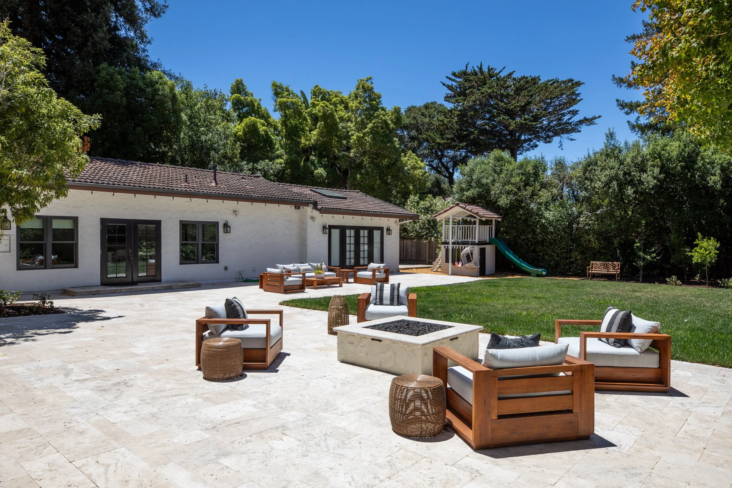A spacious backyard patio with outdoor furniture including sofas, armchairs, and side tables on a light-colored tiled surface. There is a grassy area with a wooden bench, a children's playset with a slide, and a white house with black-framed windows 