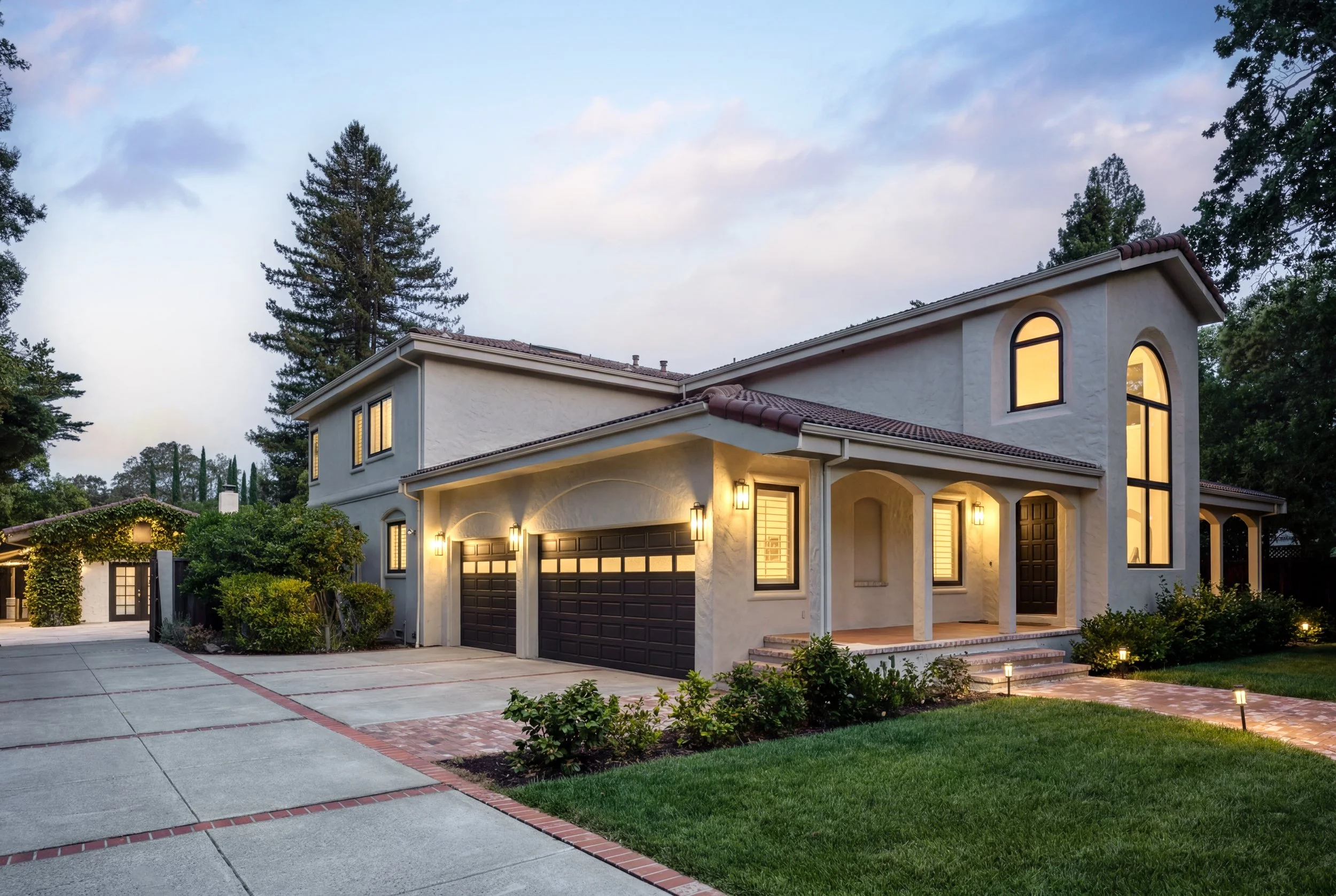 A two-story house with lit windows at dusk, featuring a garage with dark brown doors, front porch lighting, and surrounded by greenery and tall trees.