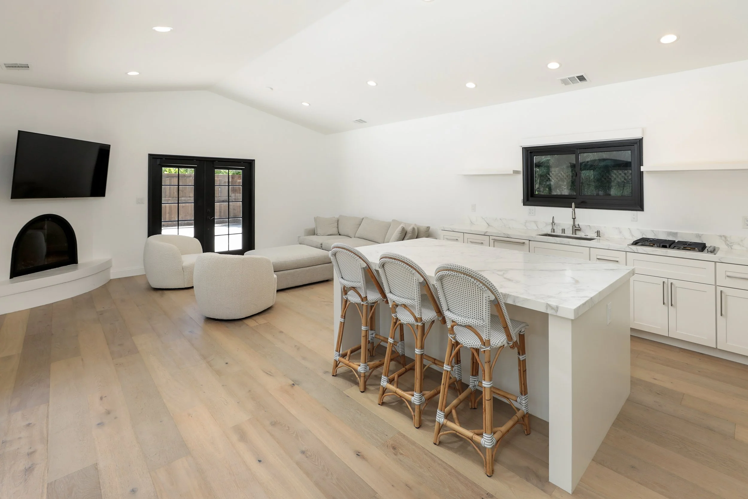 Modern living room featuring white walls and ceiling, hardwood floors, a sofa, a curved armchair, a wall-mounted TV, a fireplace, and black-framed double doors leading outside. The kitchen island has a marble countertop with four woven rattan chairs.