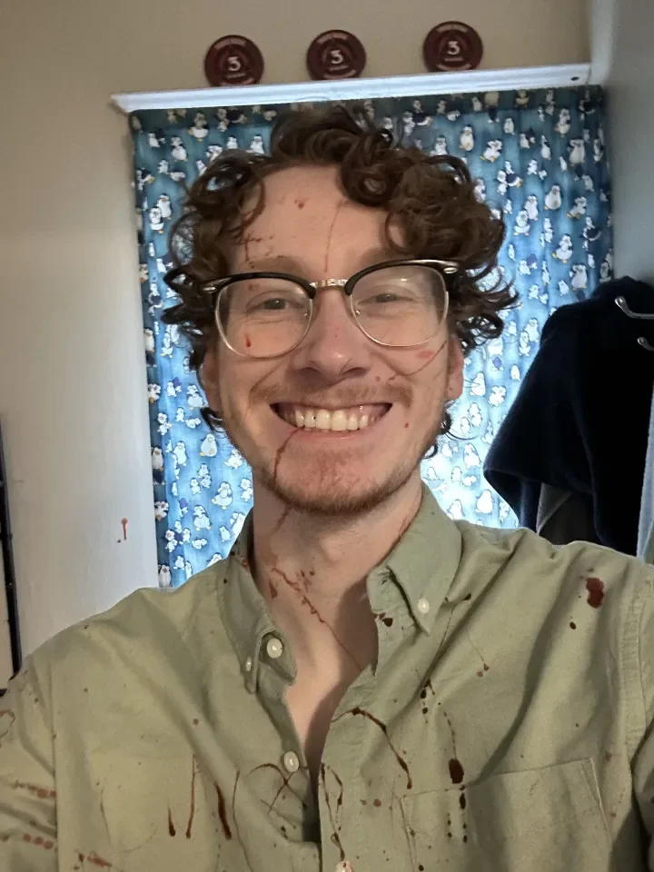 Young man with curly brown hair, glasses, and a big smile, wearing a beige button-up shirt splattered with what appears to be blood or red paint, inside a room with blue curtains featuring white snowmen or snowflake designs, and three red medals or medals hanging on the wall behind him.