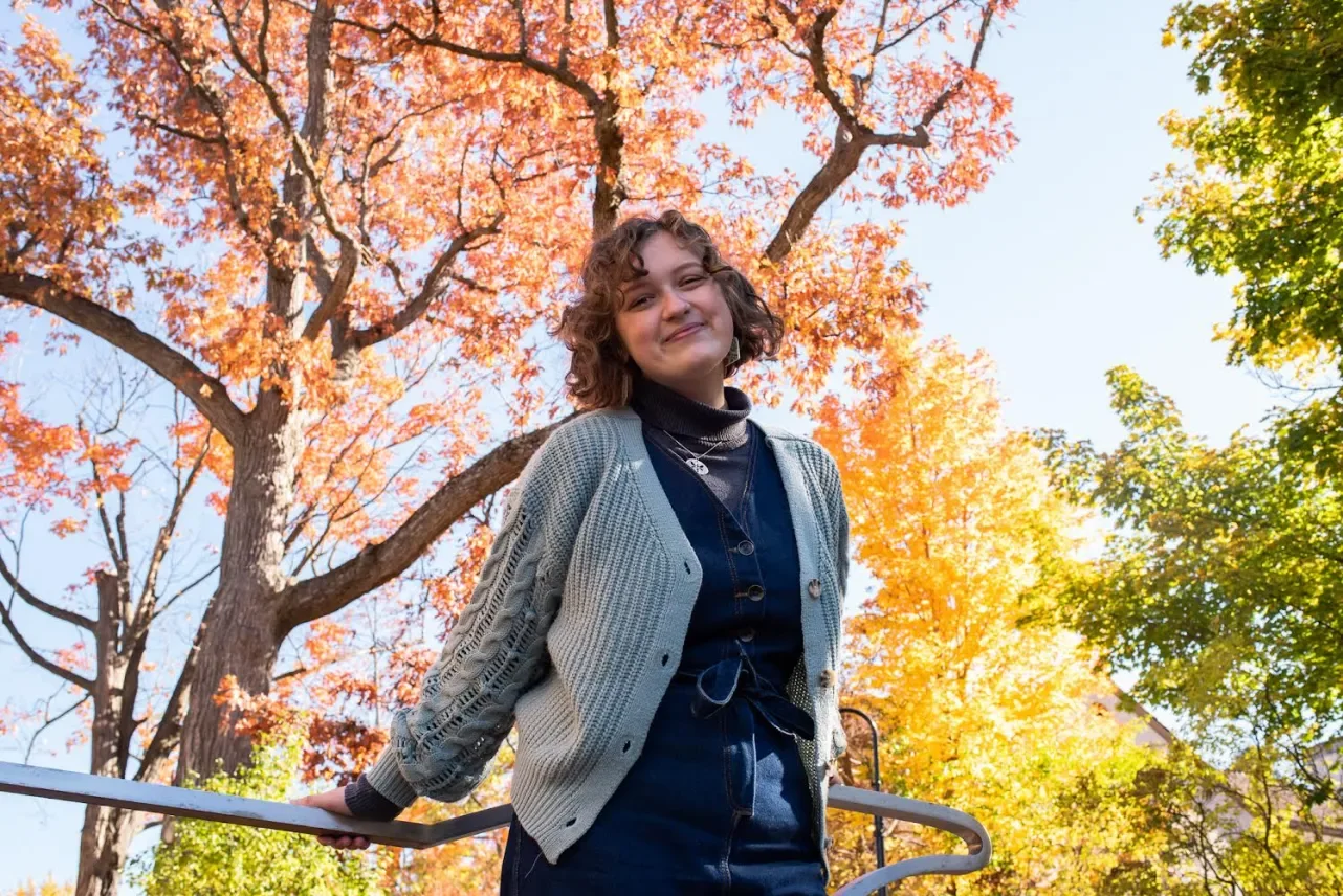 A young girl with curly hair smiling outdoors during fall, standing with her hand on a railing, surrounded by trees with orange, yellow, and green leaves.