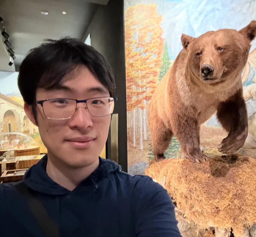 A young man with glasses takes a selfie next to a large bear statue with a painted forest background.