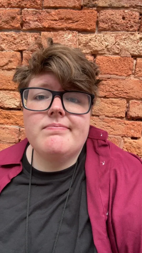 Close-up of a young person with short, wavy brown hair, wearing black glasses, a black shirt, and a maroon jacket, standing in front of a brick wall.
