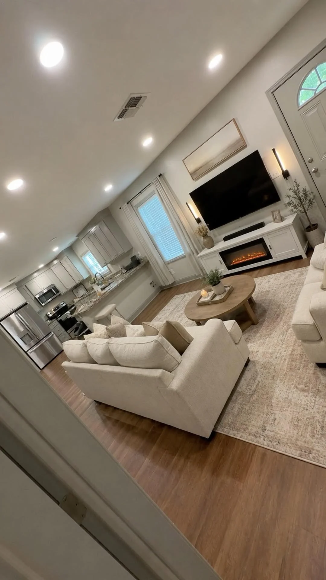A cozy living room with a beige sofa, a round wooden coffee table, a large flat-screen TV, and an electric fireplace, with windows and kitchen in the background.