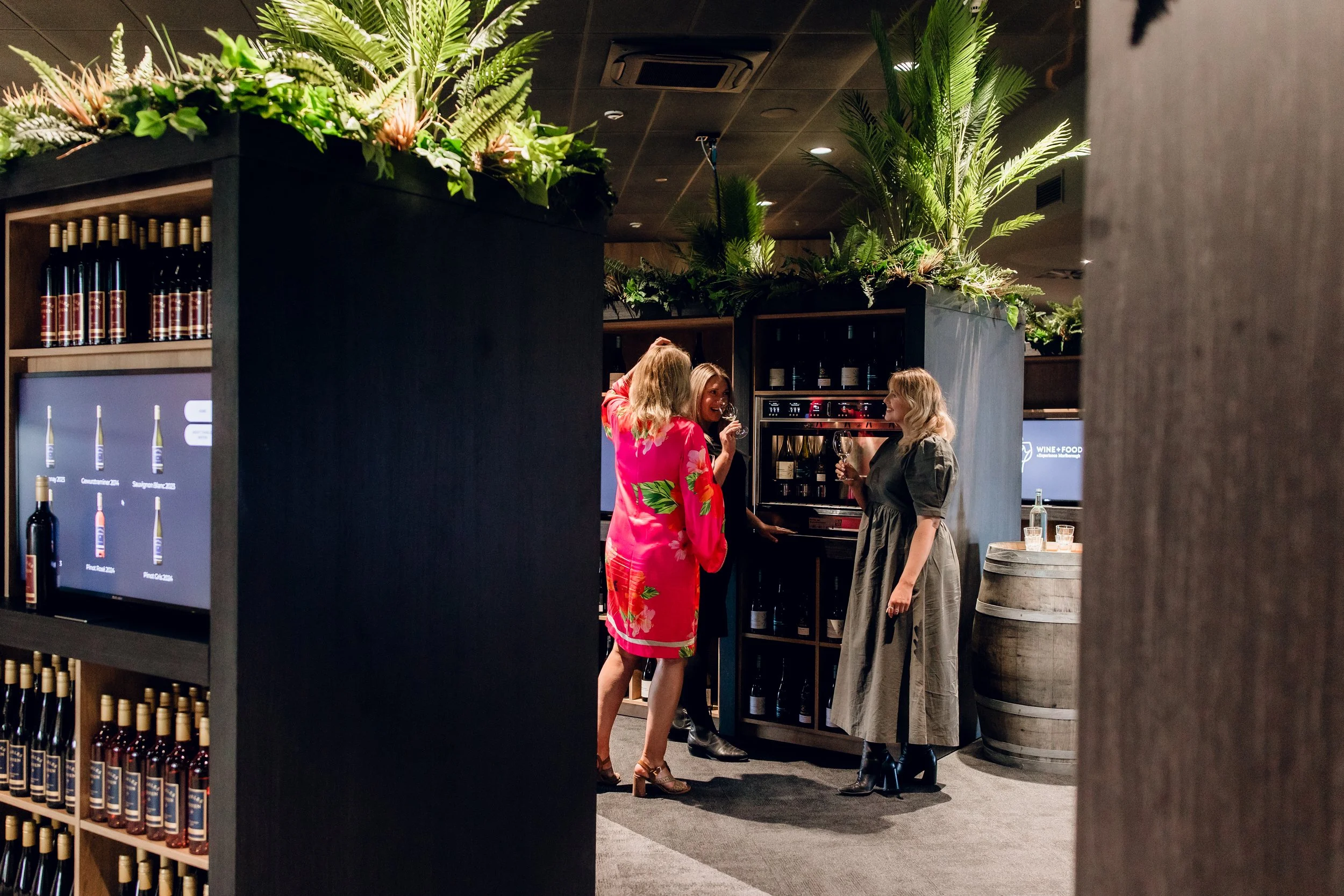 Three women stand around a wine cooler in a dimly lit wine store, chatting and drinking wine. One woman wears a red floral dress, another a gray dress, and the third a black outfit. There are large leafy plants and a wine barrel in the background.