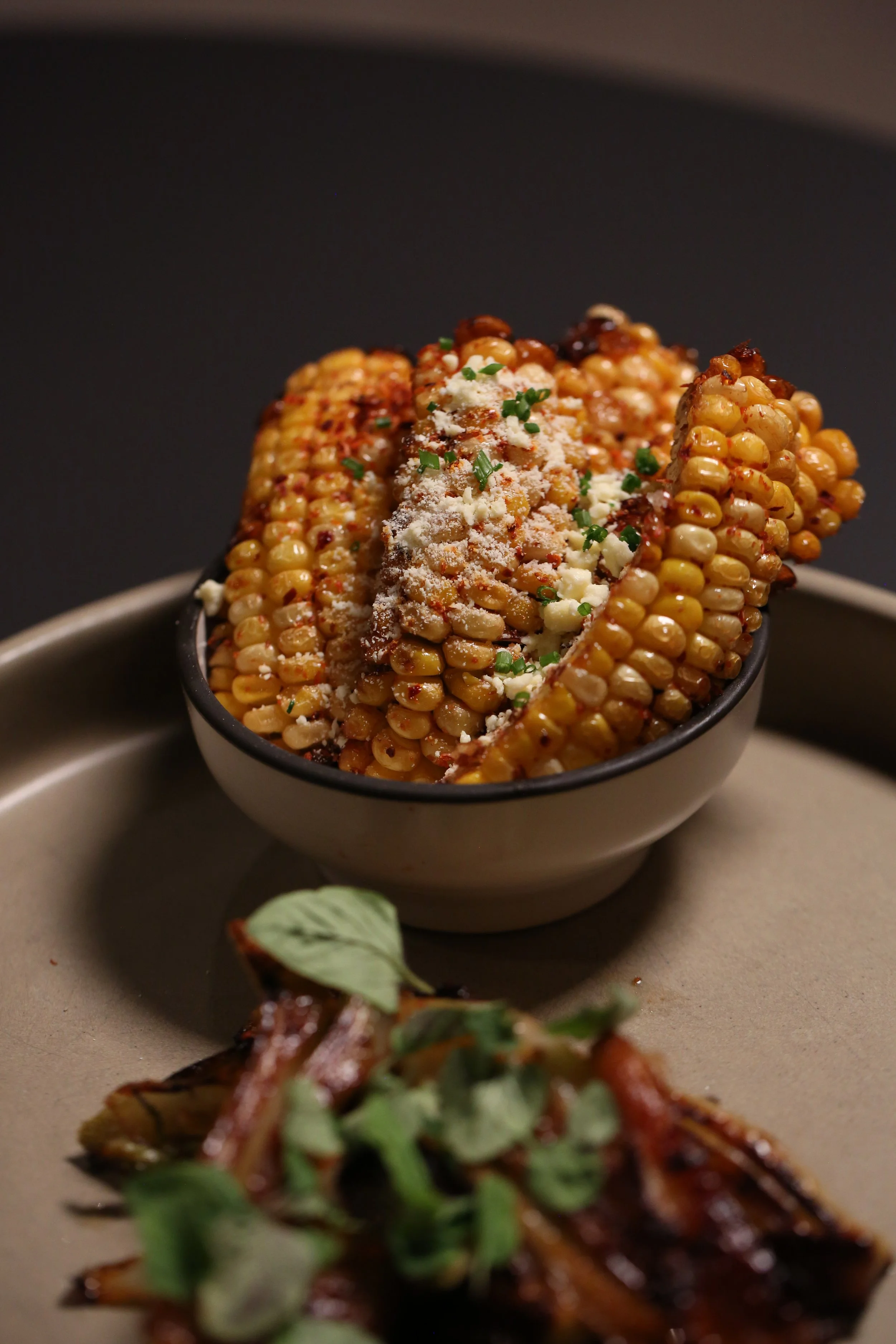 Close-up of grilled corn on the cob topped with grated cheese and chopped chives in a black bowl on a plate, with another dish partially visible in the foreground.