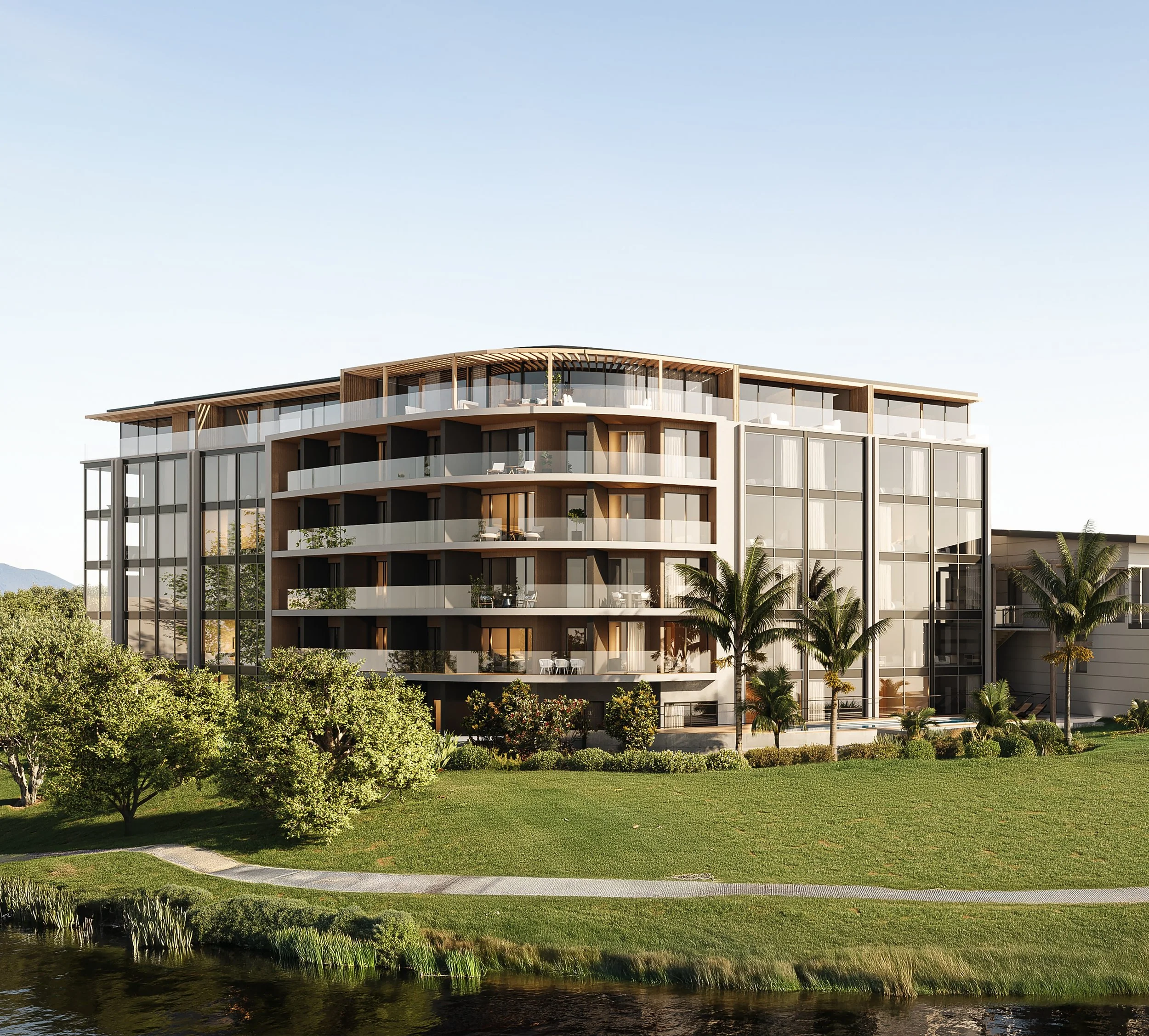 Modern multi-story building with large glass windows and balconies, surrounded by lush green trees, with a pond in the foreground and a clear blue sky overhead.