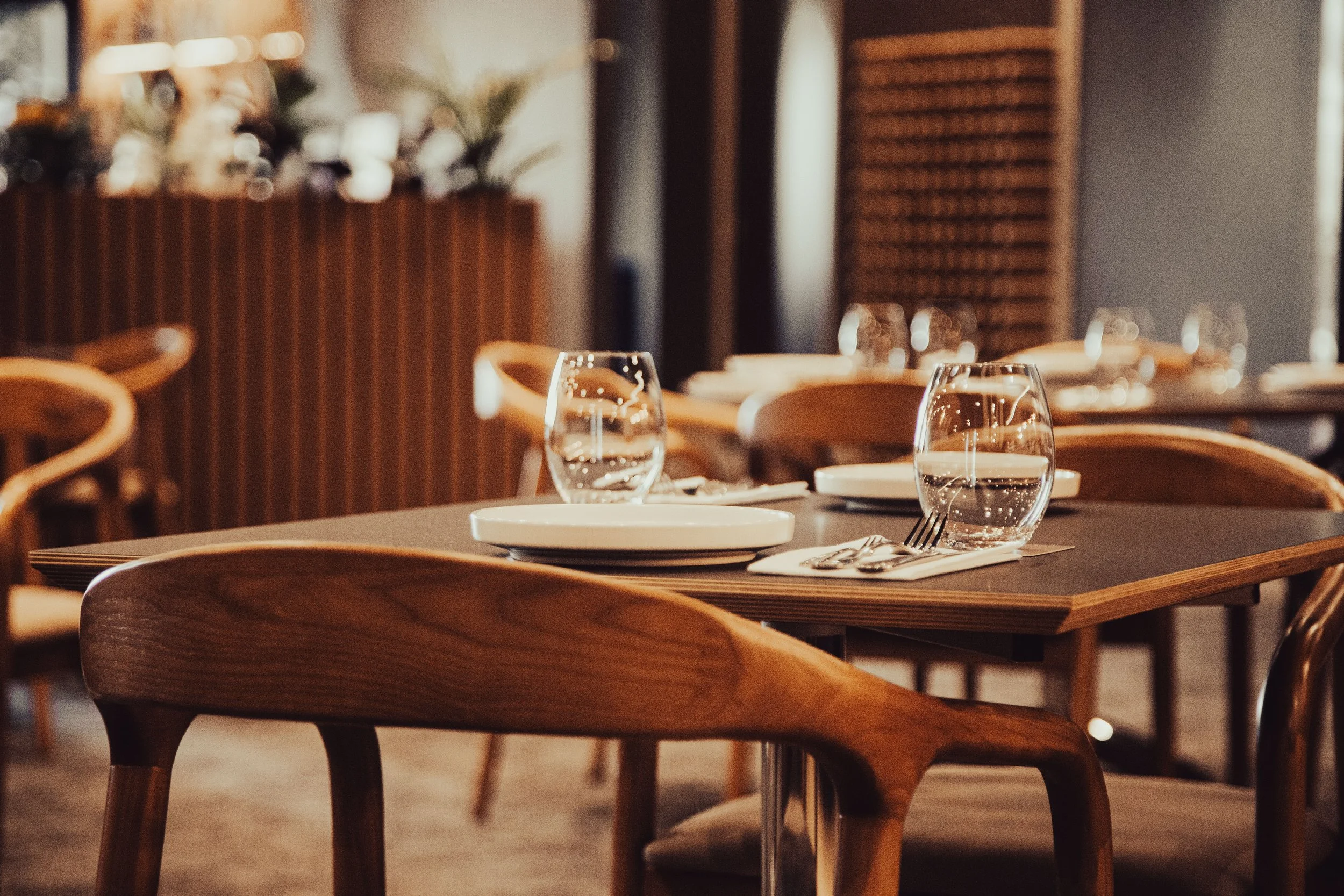 A set of wooden chairs and tables in a restaurant or cafe, with plates, drinking glasses, and silverware arranged on the tables, and warm ambient lighting.