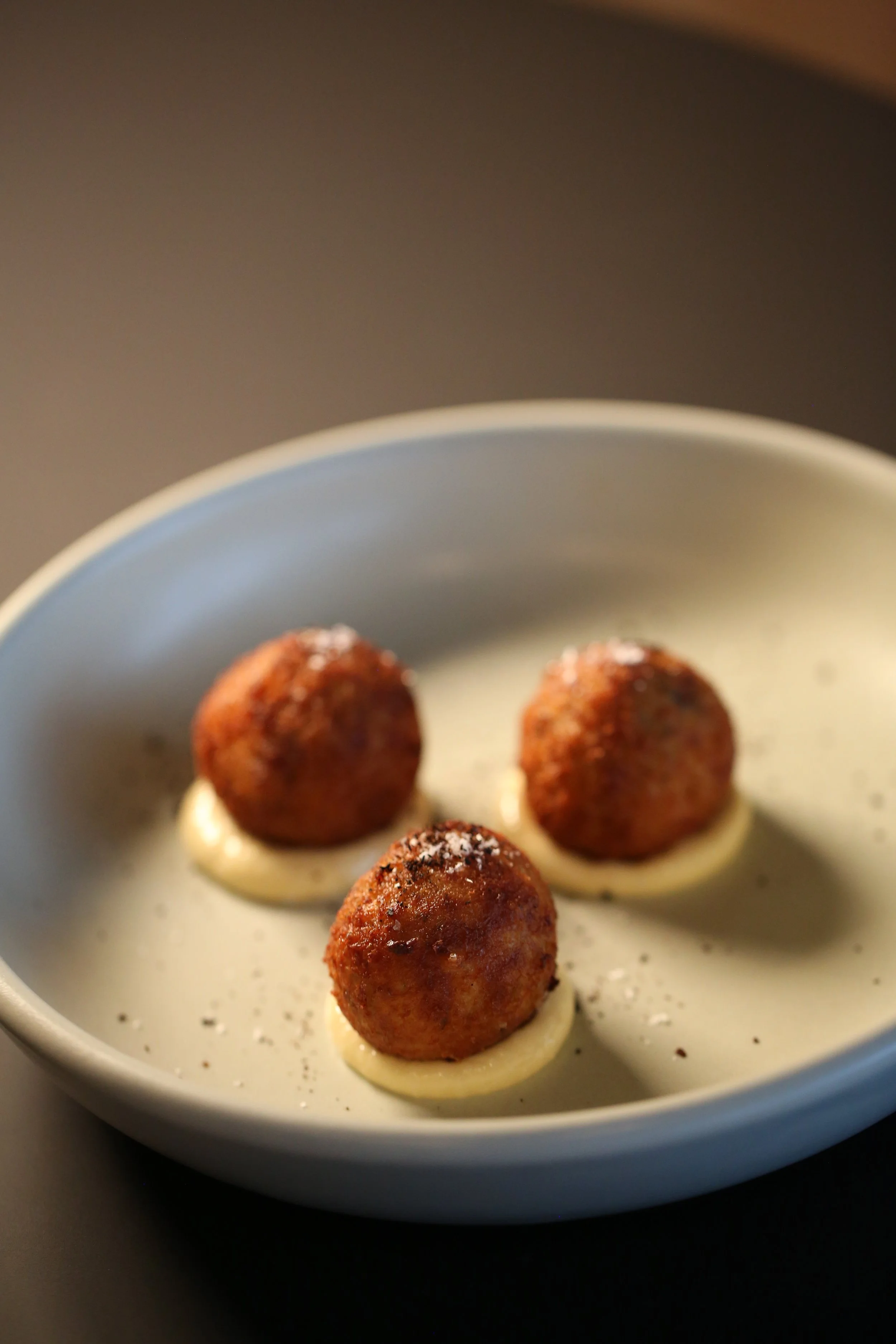 A white bowl containing five meatballs with a browned exterior, resting on a creamy white sauce base, with a light sprinkle of black pepper on the sauce.