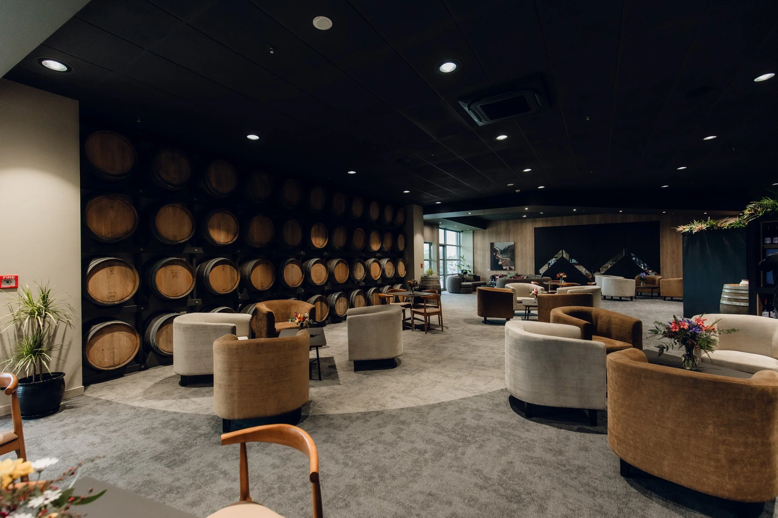 A wine cellar lounge area with round tables, comfortable chairs, a wall of wine barrels, large windows, and modern decor.
