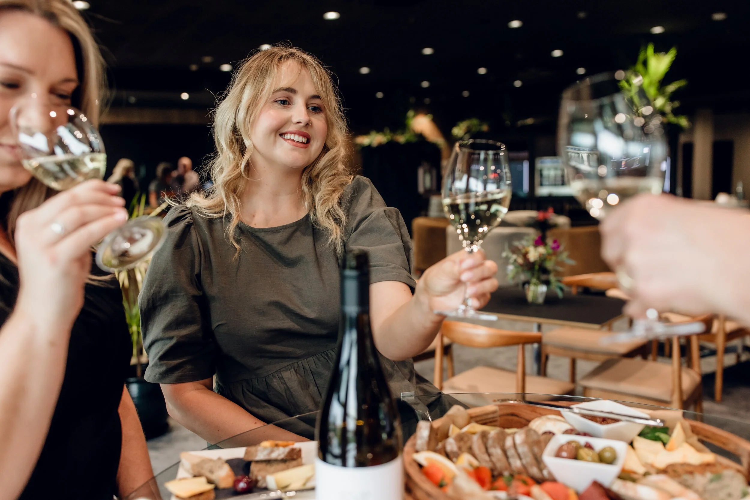 Two women sitting at a table holding glasses of white wine, smiling at each other, with appetizers and food on the table in a dimly lit restaurant or event space.