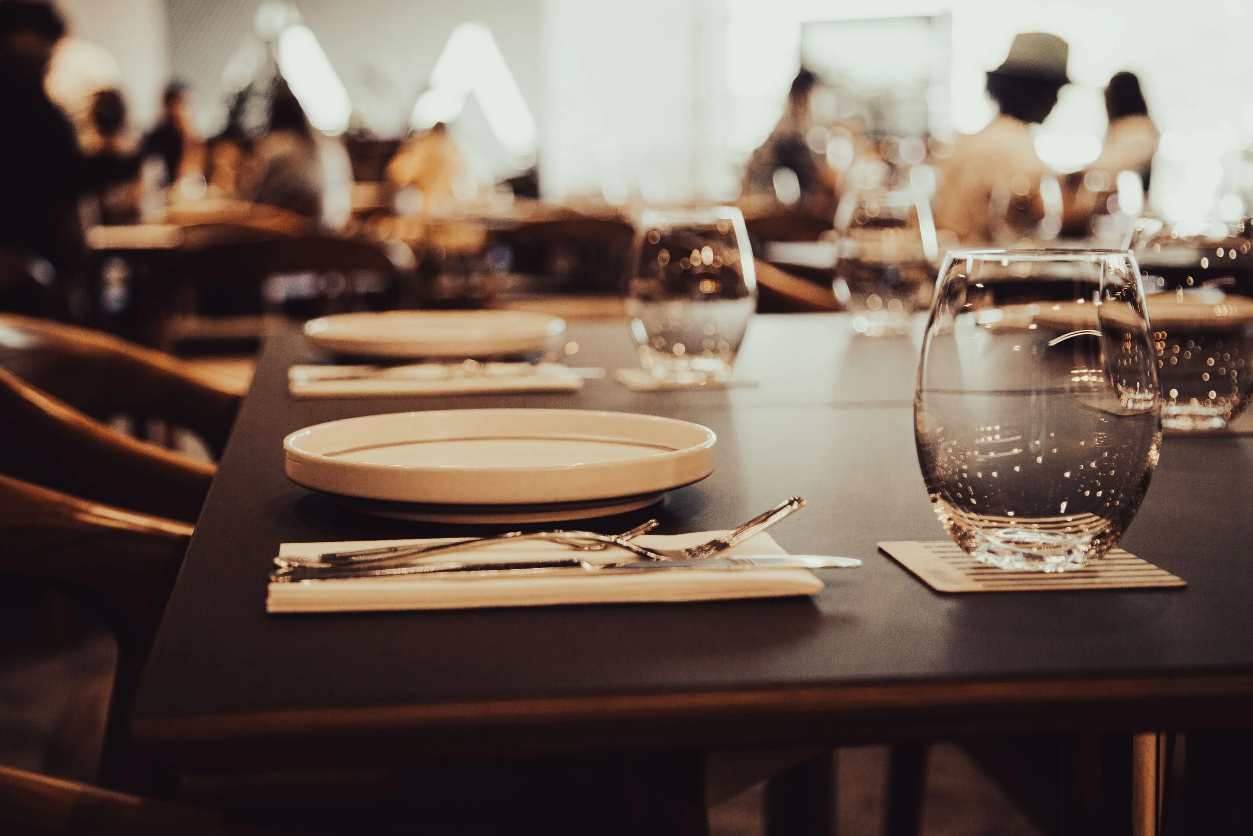 A close-up of an empty dining table set with a white plate, a drinking glass filled with water, cutlery, and a napkin on a placemat in a softly lit restaurant with blurred background of people sitting at other tables.