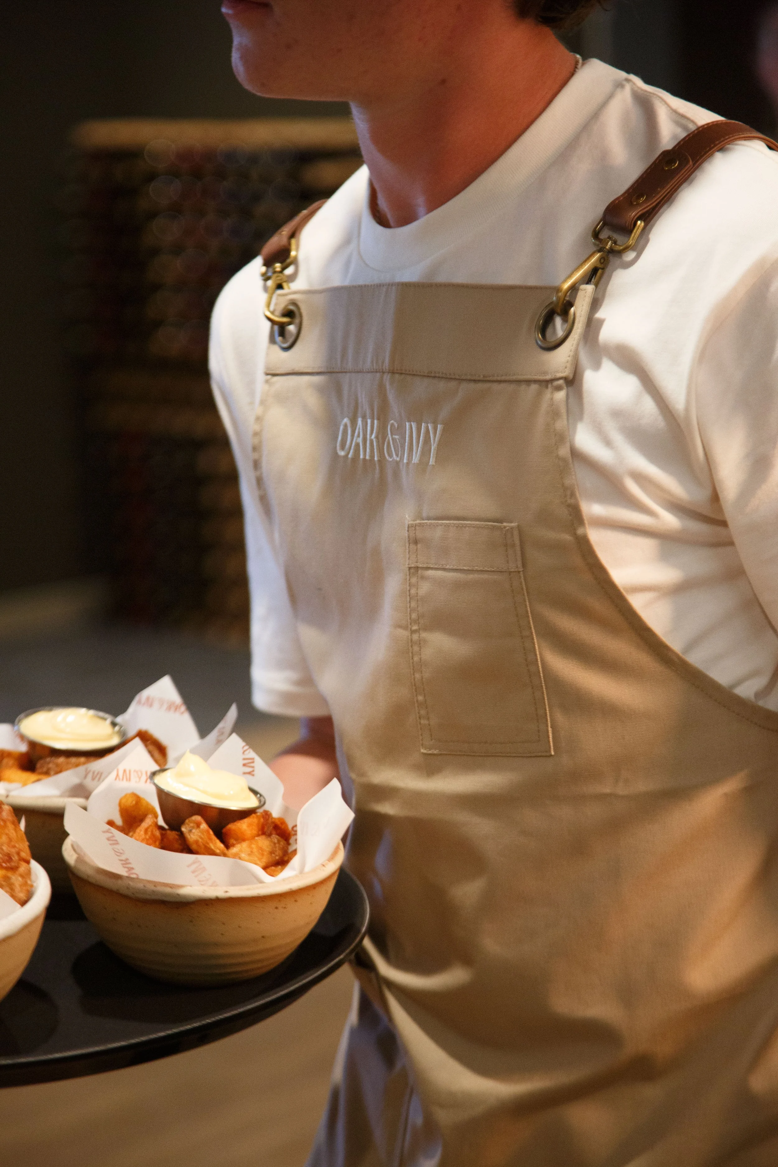 A restaurant employee wearing a beige apron with 'OAK & IVY' embroidered on it carries a tray of fried appetizers with dipping sauces.