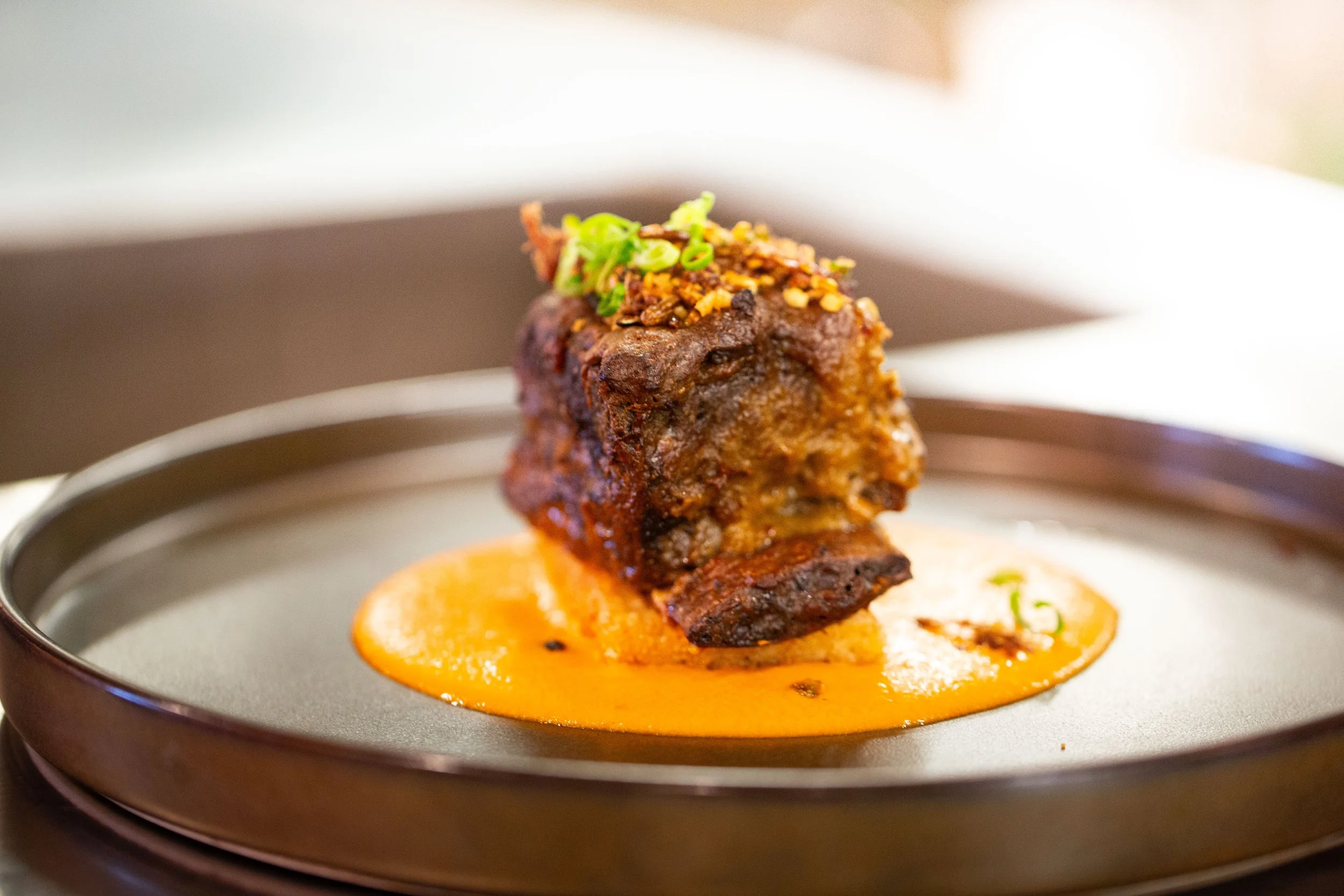 Close-up of a piece of cooked meat with diced vegetables and green onions on top, served on a bed of orange sauce on a plate.