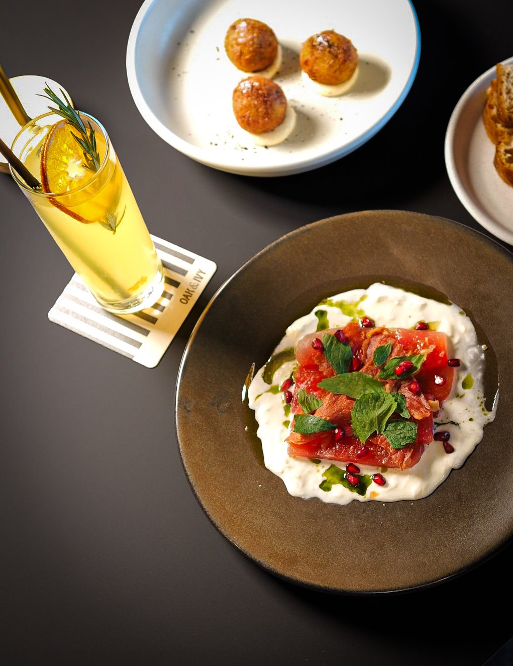 A plate of Mediterranean-style dish featuring sliced tomatoes, fresh herbs, pomegranate seeds, and a creamy white sauce on a dark plate. A glass of lemon-infused beverage garnished with lemon slices, herbs, and a straw is nearby. In the background, t