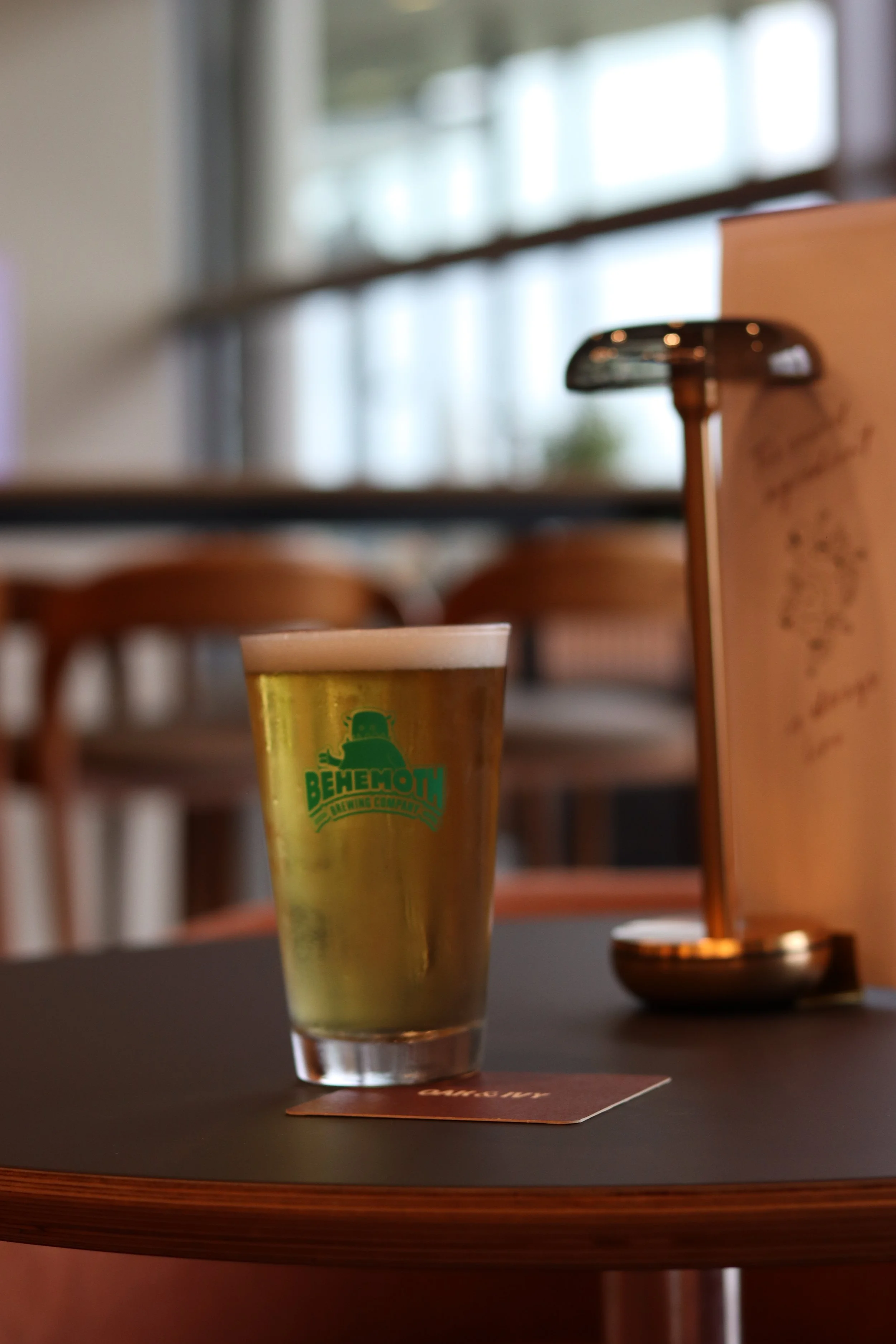 Close-up of a glass with yellow-green beer with a logo that reads 'Behemoth Brewing Company,' placed on a dark table in a well-lit indoor setting.