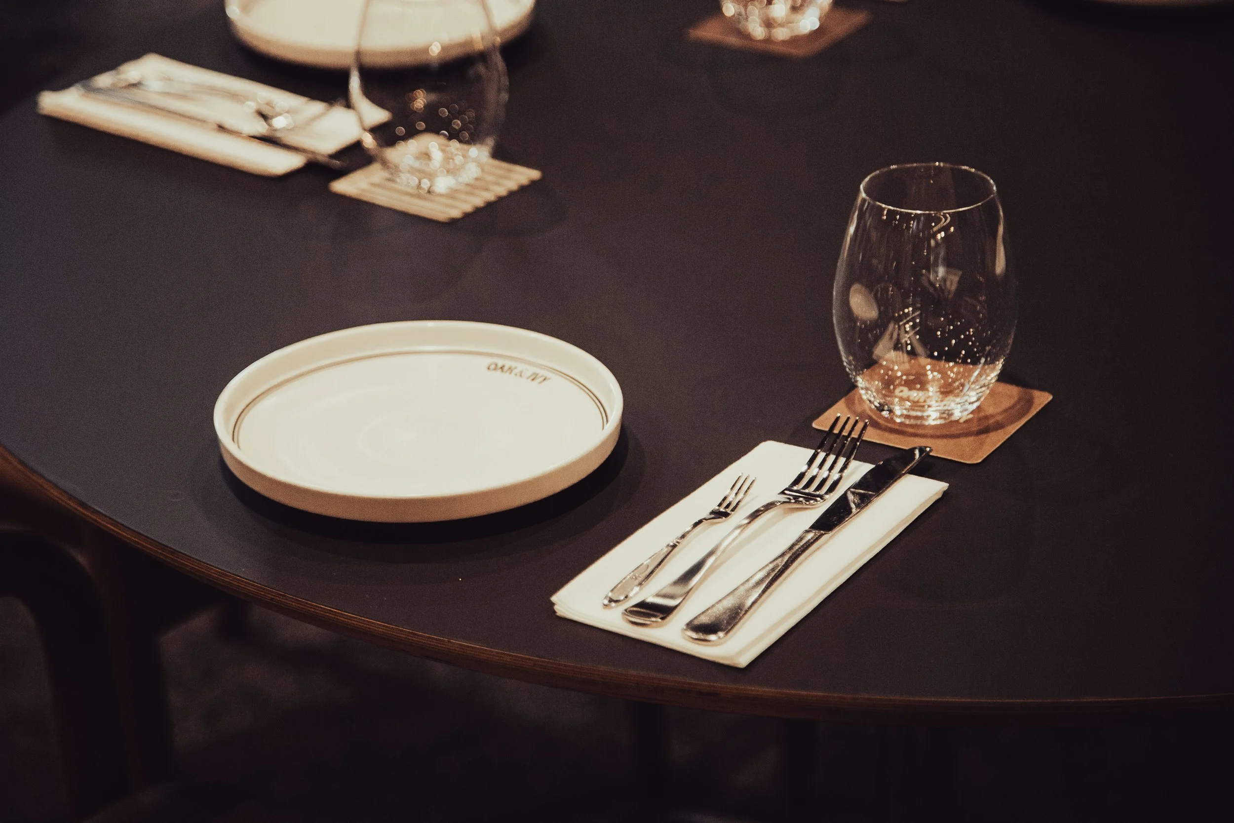 A formal table setting on a dark tablecloth with a white plate, clear water glass on a wooden coaster, and neatly arranged silverware on a folded white napkin.