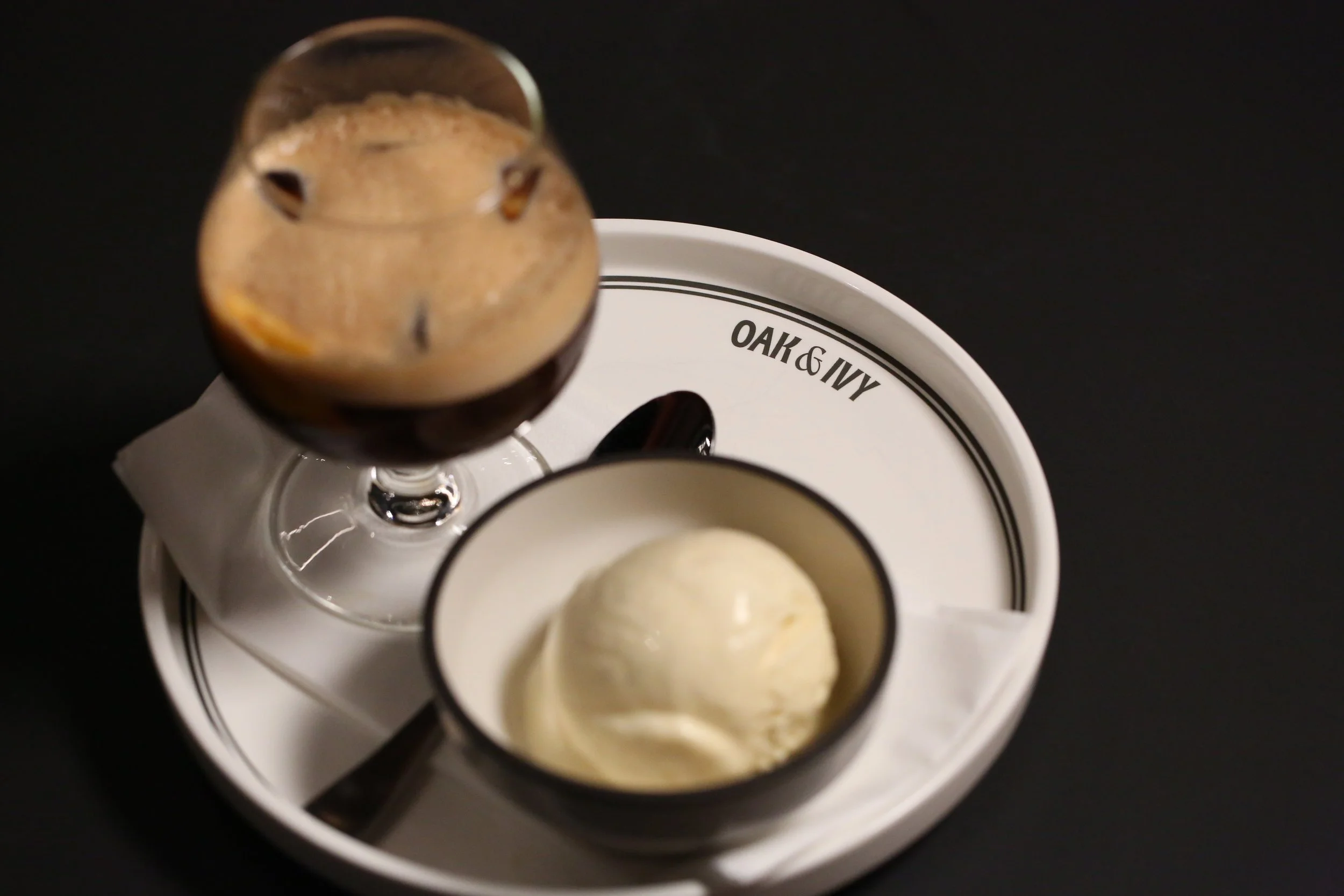 A white Oak & Ivy plate holding a glass of iced coffee and a small dish of vanilla ice cream, with a black background.