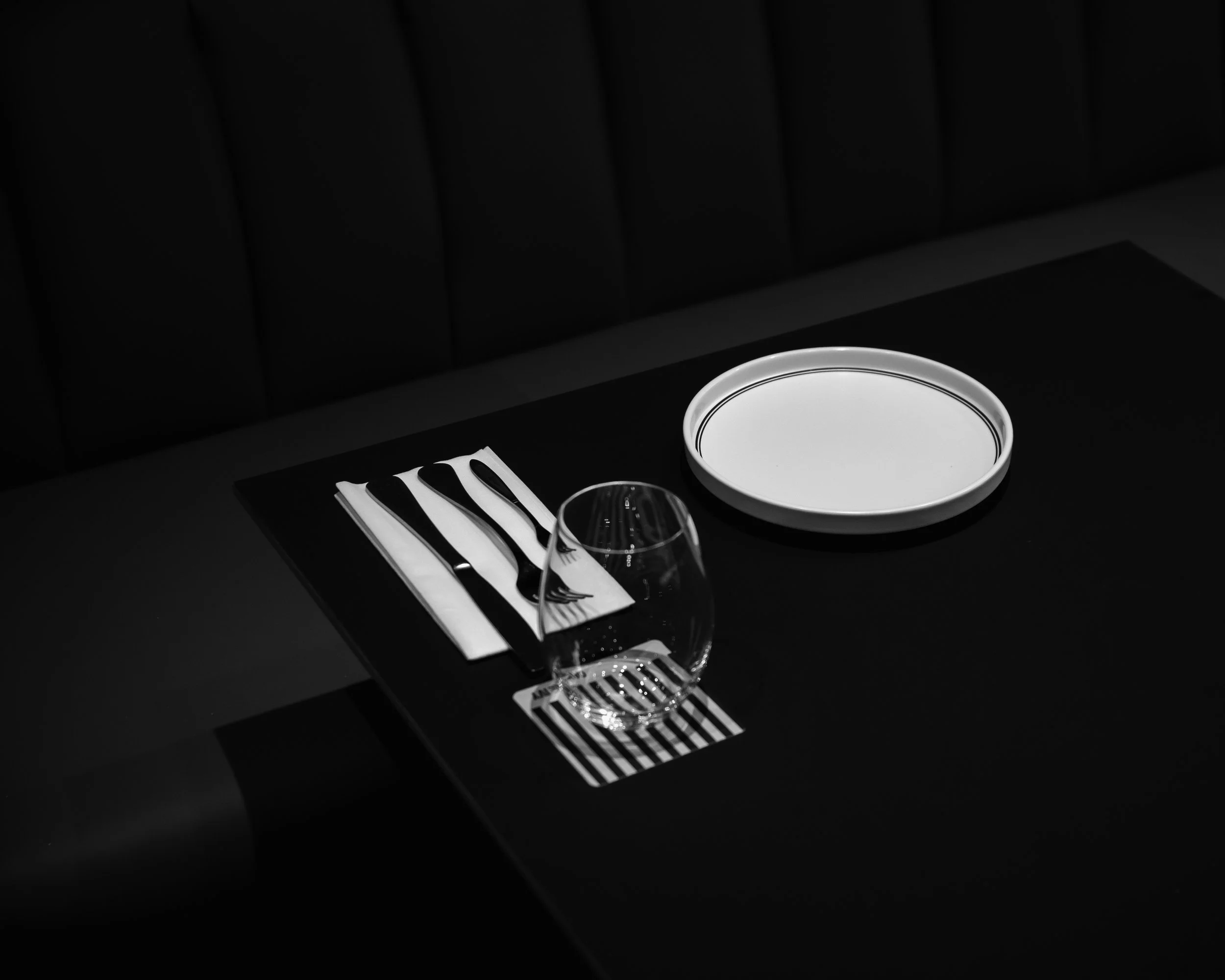 A black table setting with a white plate, a fork, a knife, a spoon, and a clear wine glass, with a black and white striped napkin underneath.