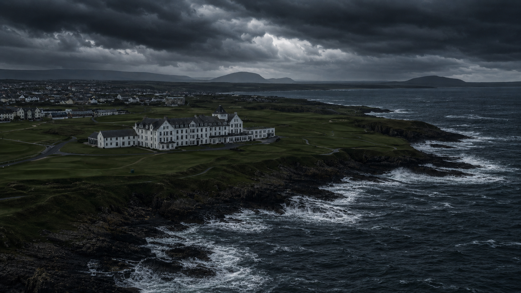A large white hotel on a rocky coastline under a dark, cloudy sky, with waves crashing against the shore in the foreground.