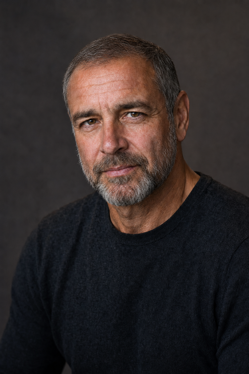 George 'Seoirse' Ryan -Close-up portrait of a middle-aged man with gray hair and a beard, wearing a black shirt, against a dark gray background.