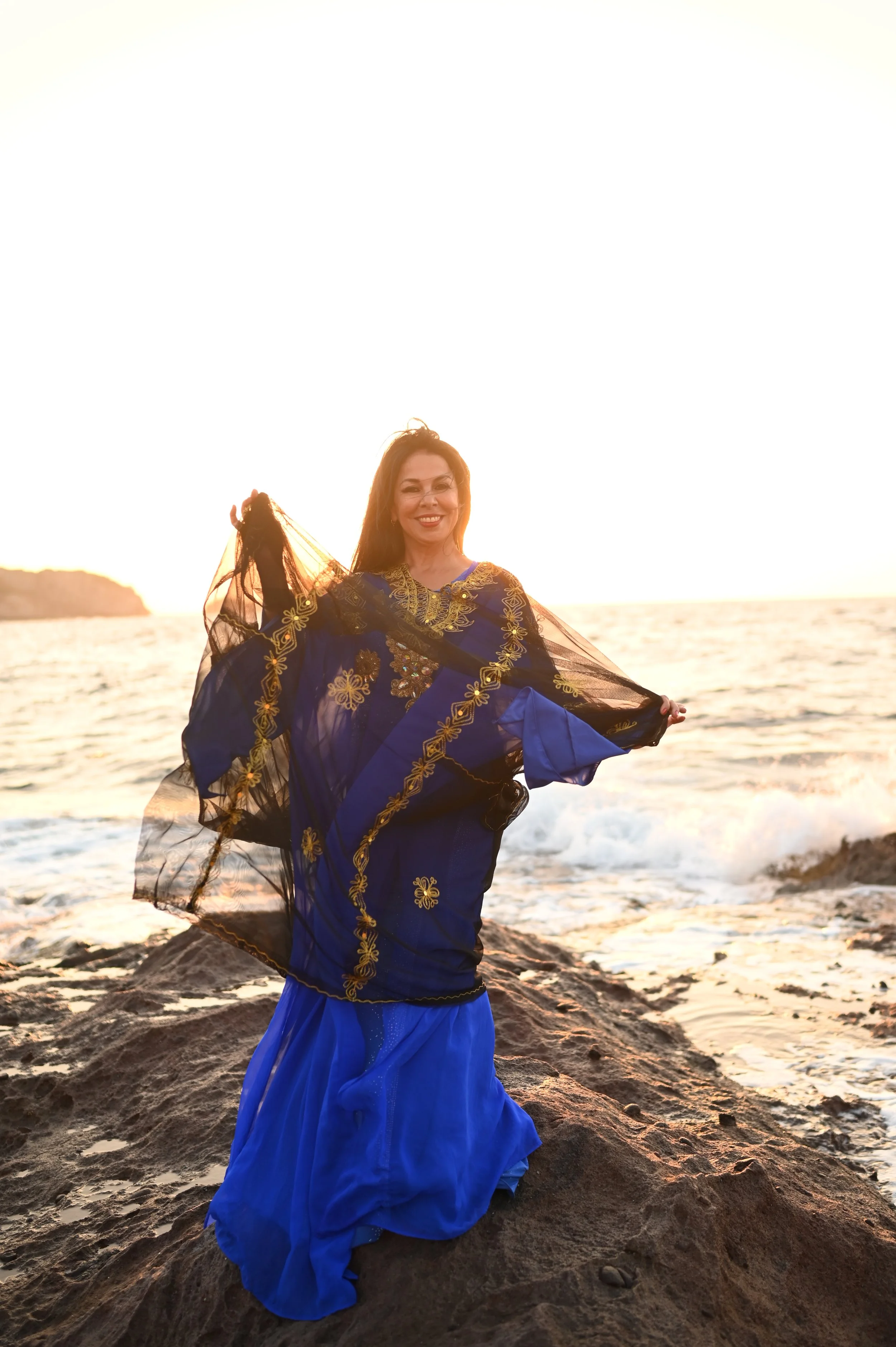 Woman in traditional blue and black dress with gold embroidery standing on rocky beach at sunset with ocean in the background.