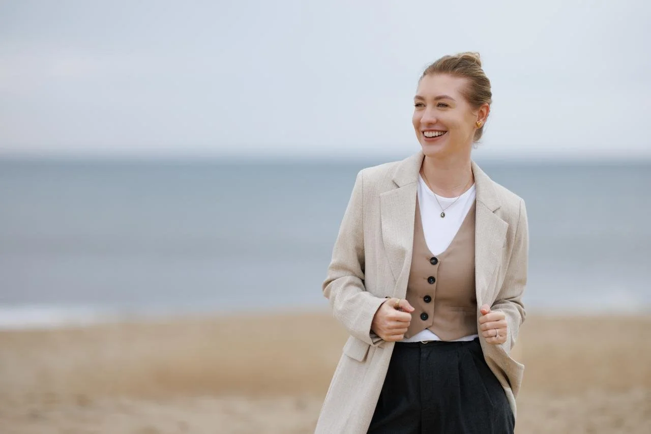 A woman smiling and walking on the beach, dressed in a beige blazer over a beige vest, white shirt, and dark pants, with the ocean in the background.