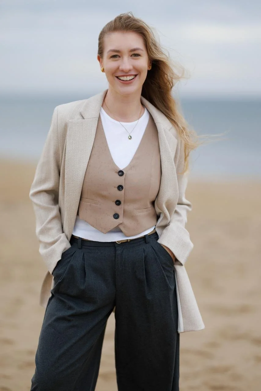 A woman with long blonde hair smiling on a beach, dressed in a beige blazer over a white shirt, a tan vest, and black pants.