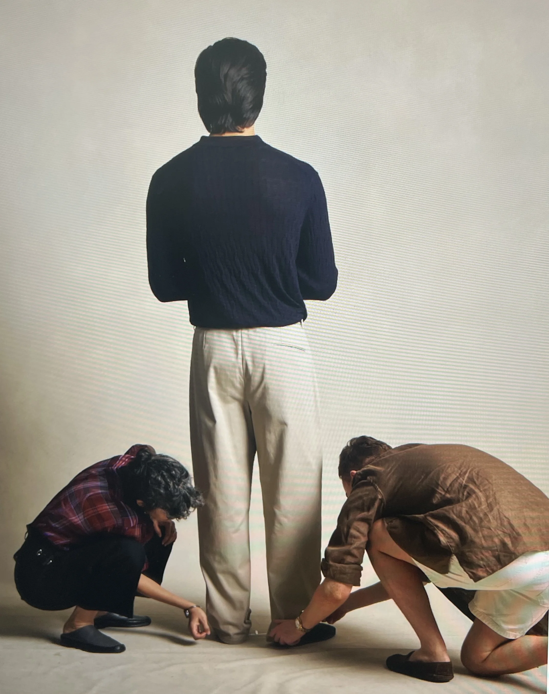 People working together on a project, bending down and holding objects on the floor, with one person standing and facing away from the camera against a plain wall.
