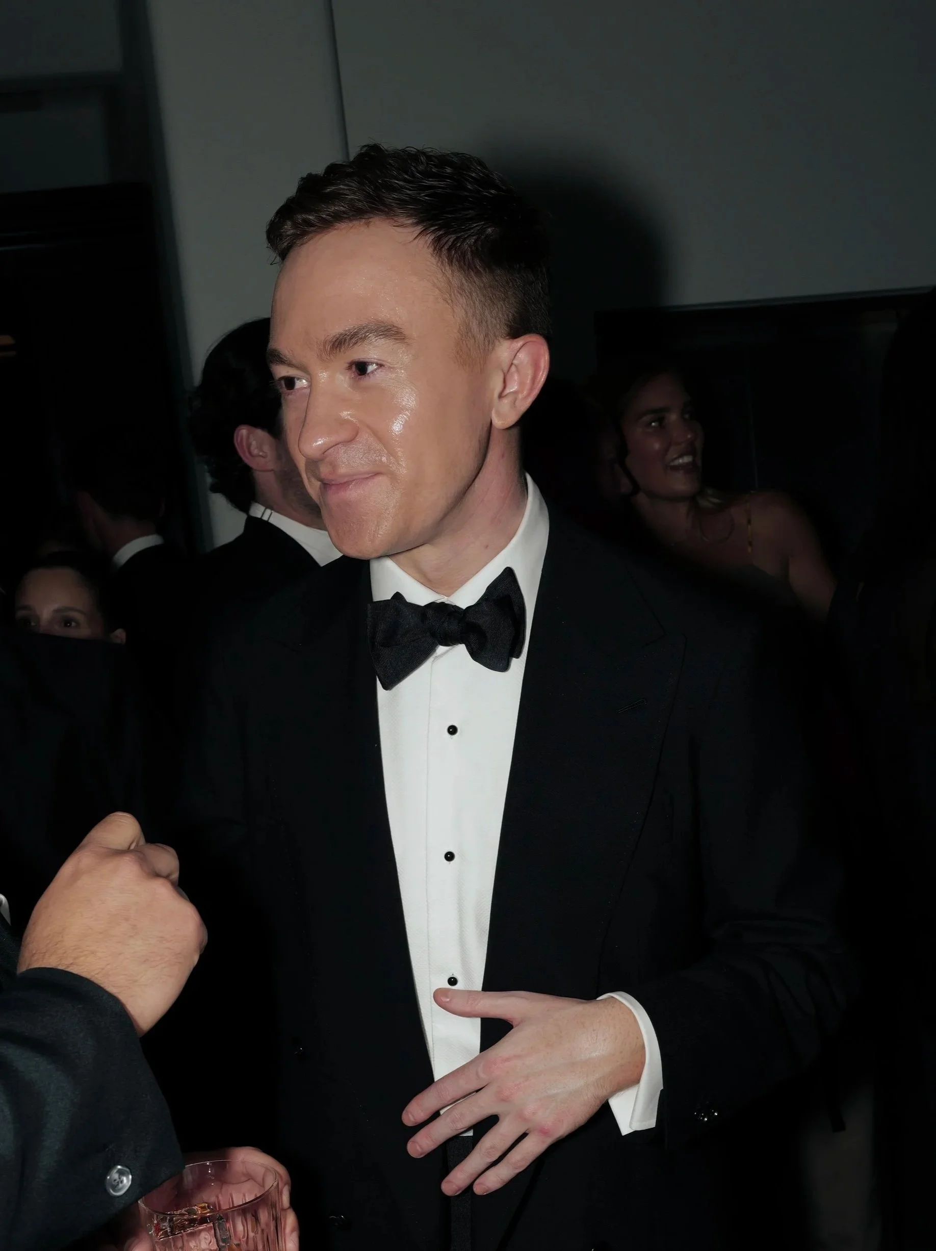A man in a tuxedo and bowtie adjusting his cuff at a social event, with people and dim lighting in the background.