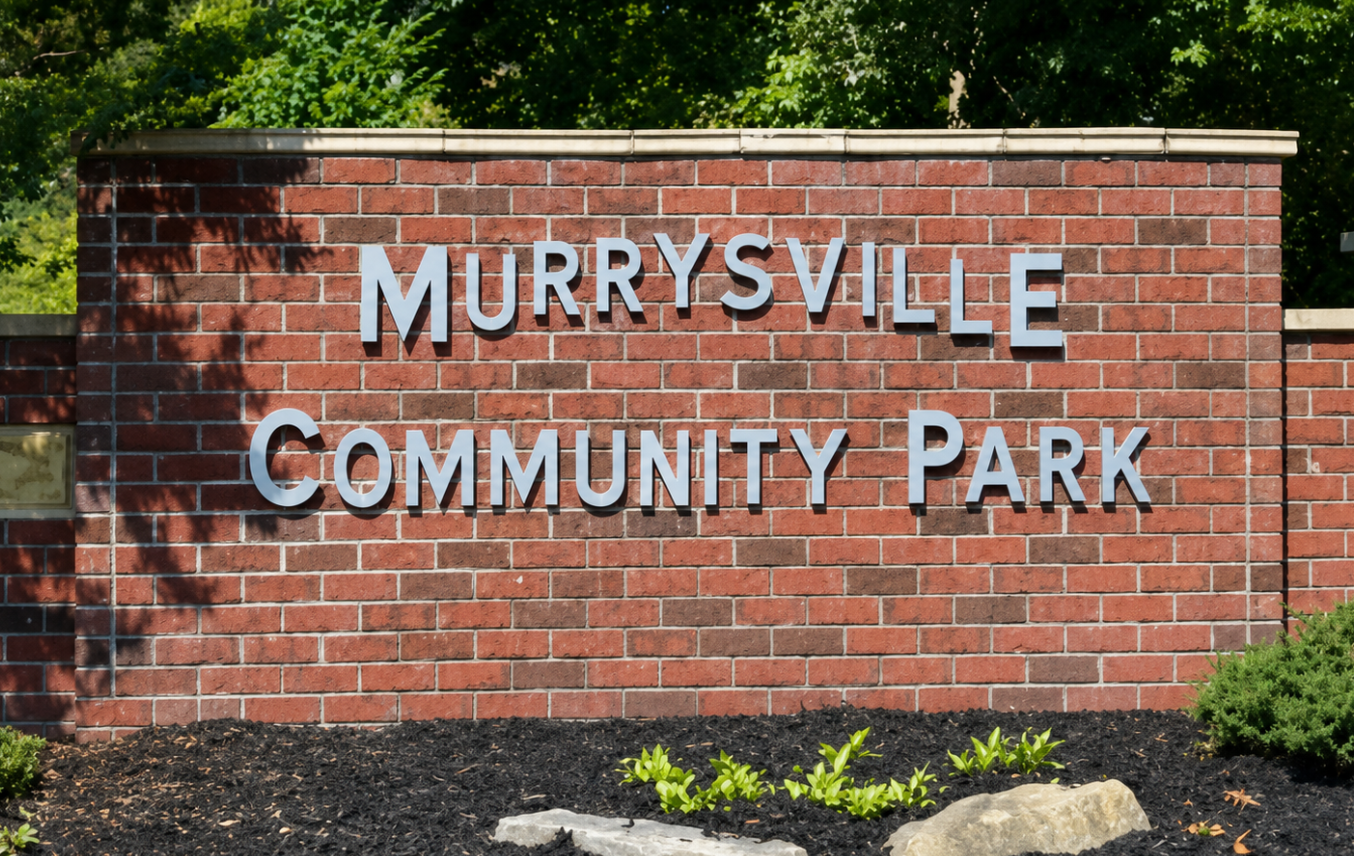 A brick sign with large metal letters reads 'MURRYSVILLE COMMUNITY PARK.' There are trees behind the sign and some small shrubs and rocks in front of it.