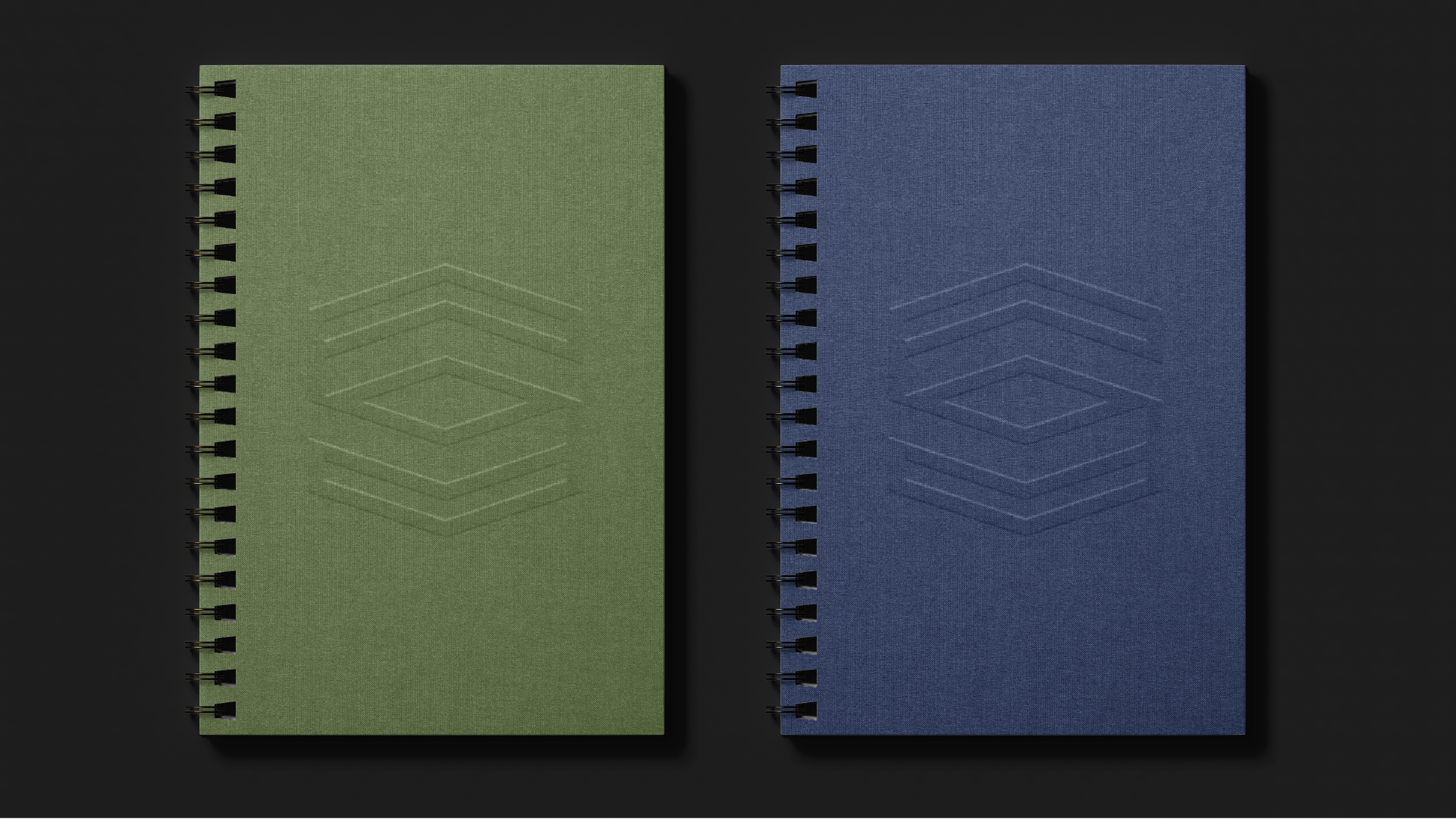 Two spiral-bound notebooks, one green and one blue, lying flat on a dark surface with subtle geometric patterns embossed on their covers.