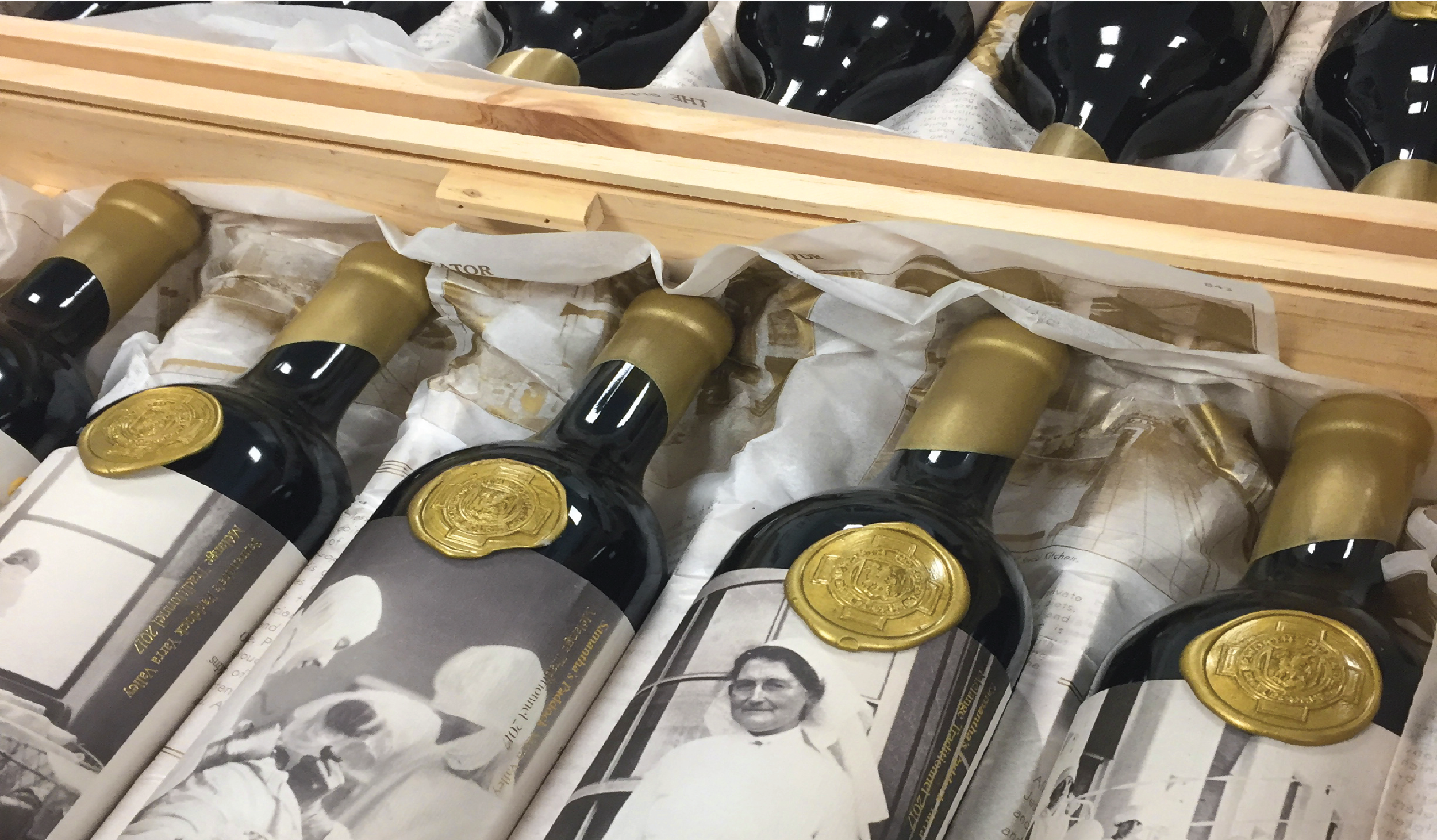 Bottles of sparkling wine with gold foil and black labels, wrapped in newspaper, and a piece of wood on top.