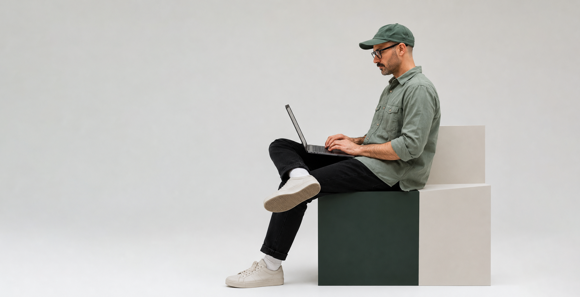A man wearing glasses, a green cap, a green shirt, black pants, and white sneakers, sitting on a geometric block while working on a laptop against a plain white background.