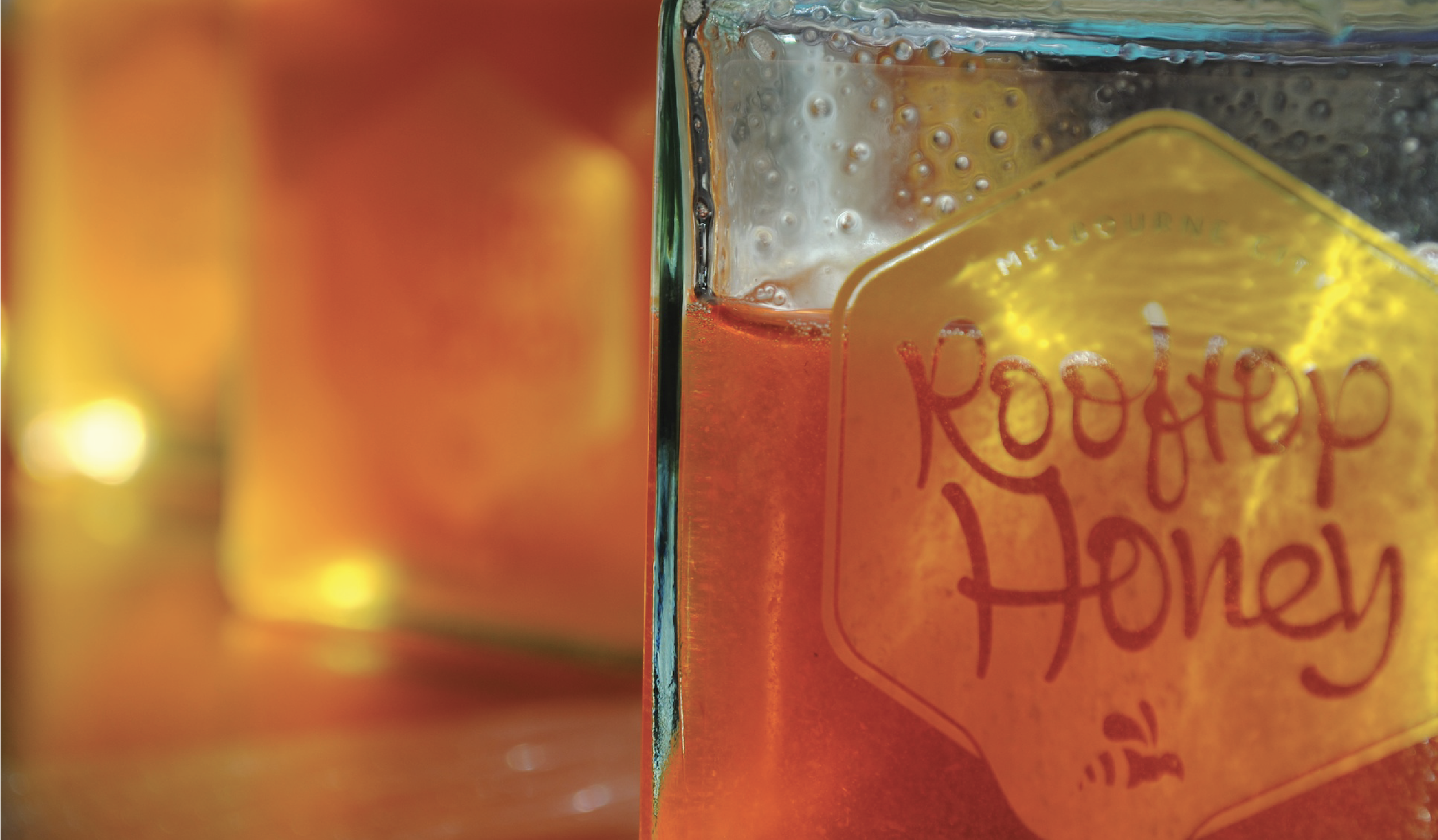 Close-up of a glass with bubbles and a partially visible orange drink label that reads "Rooted" with a handwritten style. The background is blurred with warm colors.