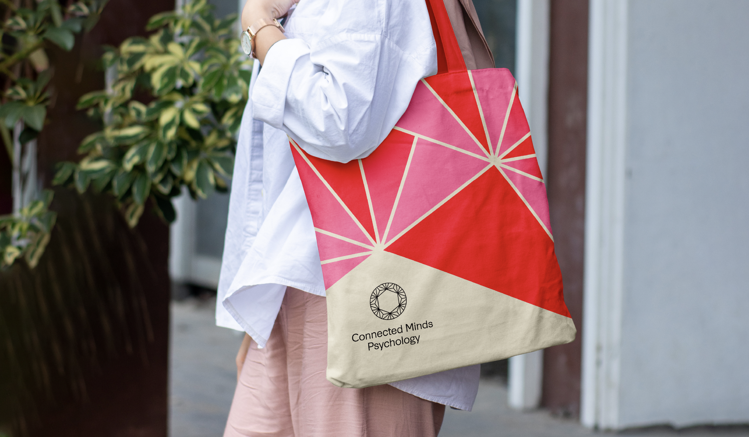 Person carrying a beige tote bag with a geometric pink, red, and beige pattern and the words "Connected Minds Psychology" and a circular logo on the bag, wearing a white shirt with rolled-up sleeves and pink pants, outside near foliage.