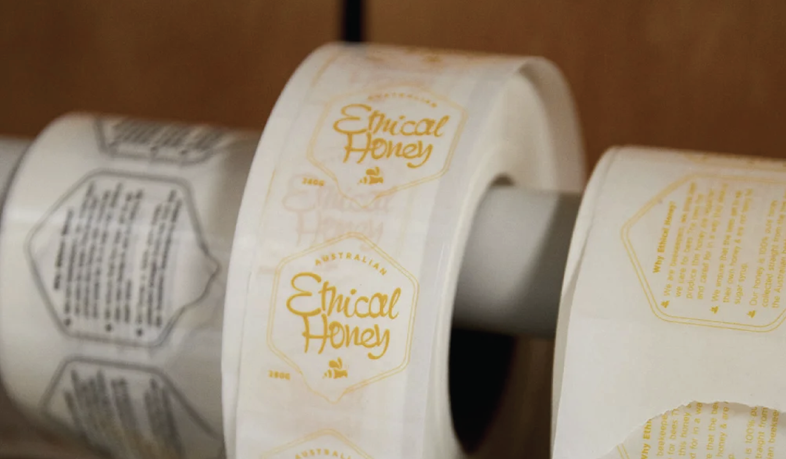 Close-up of a roll of labels with the words 'Ethical Honey' printed in yellow, featuring a honey jar illustration, on a shelf with similar products.