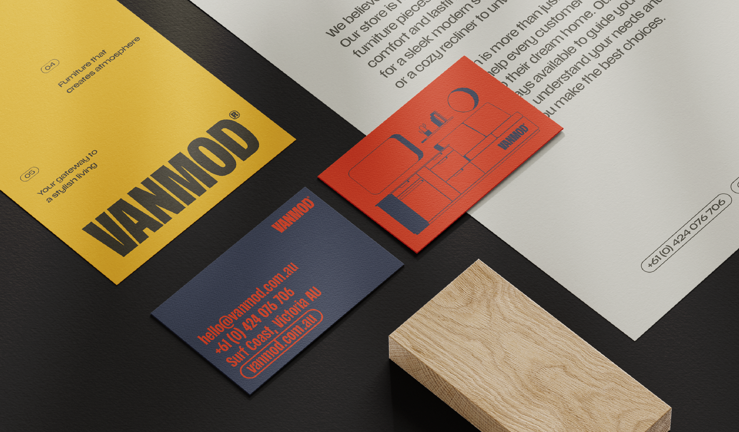 Close-up of business cards and printed material on a desk, including a yellow card with black text, a red card with blue outline of furniture, and a navy blue card with red text. A small wooden block is also visible.