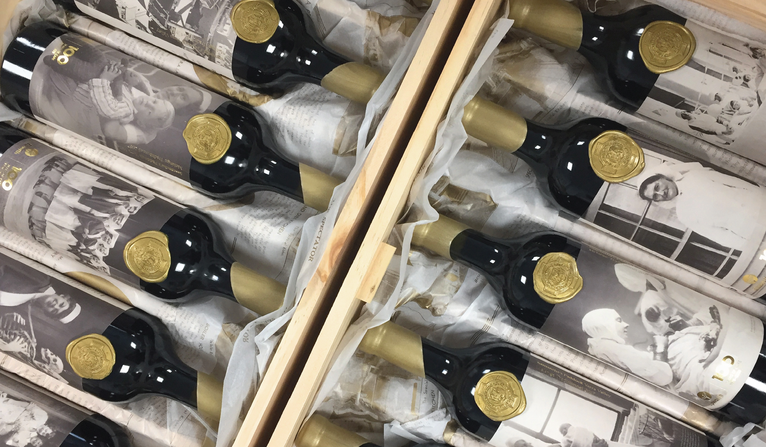 Bottles of champagne wrapped in newspaper with black and gold labels and gold foil caps, arranged in crates with crumpled newspaper packaging.